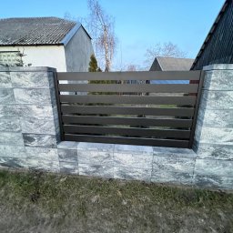 Taras Malena - Nowoczesne ogrodzenie z szarych, betonowych bloczków i poziomych, metalowych przęseł na tle domów i błękitnego nieba.