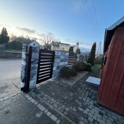 Taras Malena - Nowoczesne ogrodzenie panelowe z elementami imitującymi kamień i poziomymi przęsłami w kolorze grafitowym, widoczne z perspektywy chodnika wyłożonego kostką brukową, w tle budynki mieszkalne...