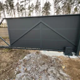 Przesuwna brama wjazdowa w kolorze grafitowym z widocznym napędem automatycznym, zamontowana na betonowym fundamencie, w tle drzewa.