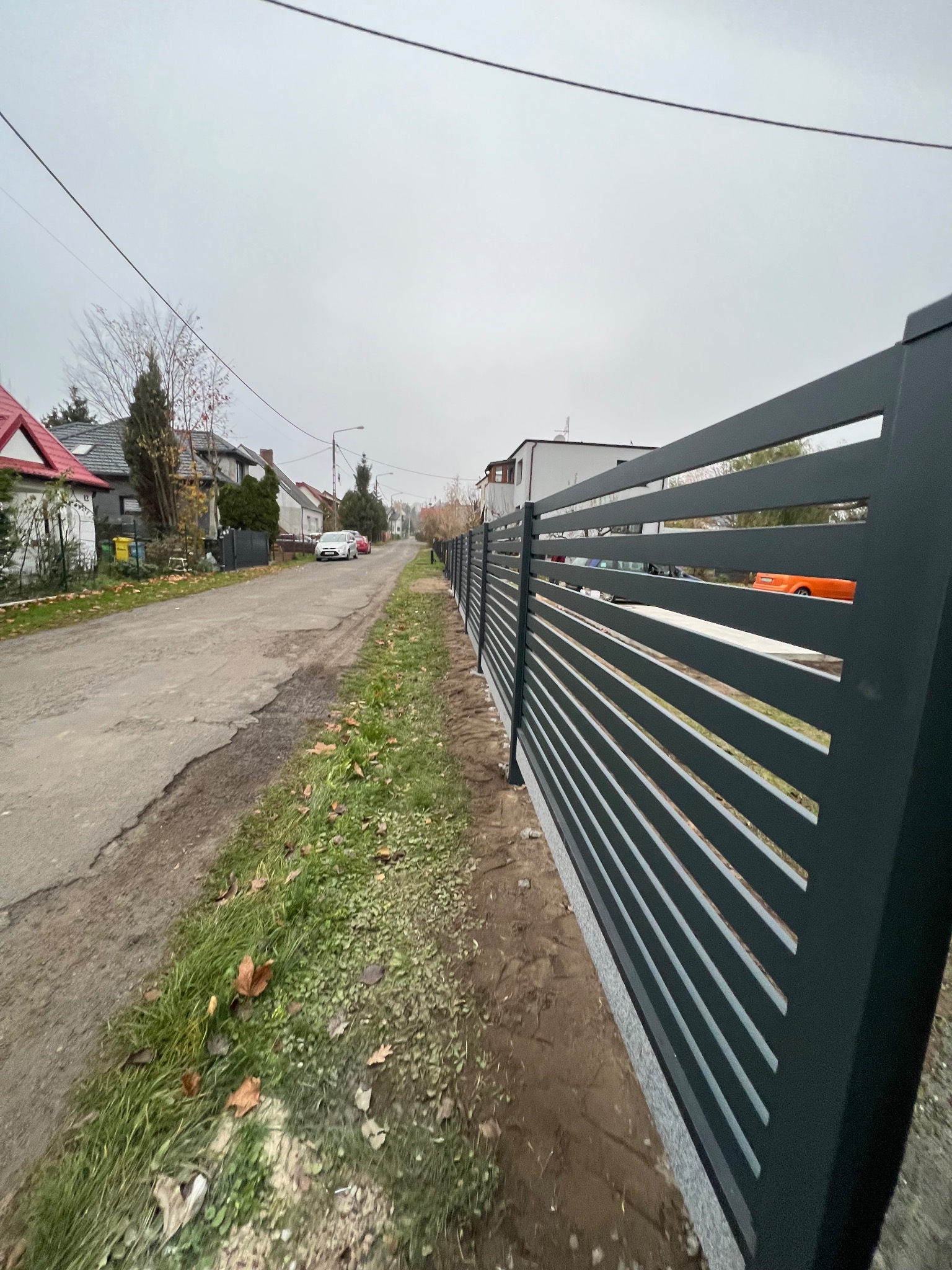 Nowoczesne, poziome, szare ogrodzenie panelowe z widokiem na ulicę i domy w tle, w pochmurny dzień.