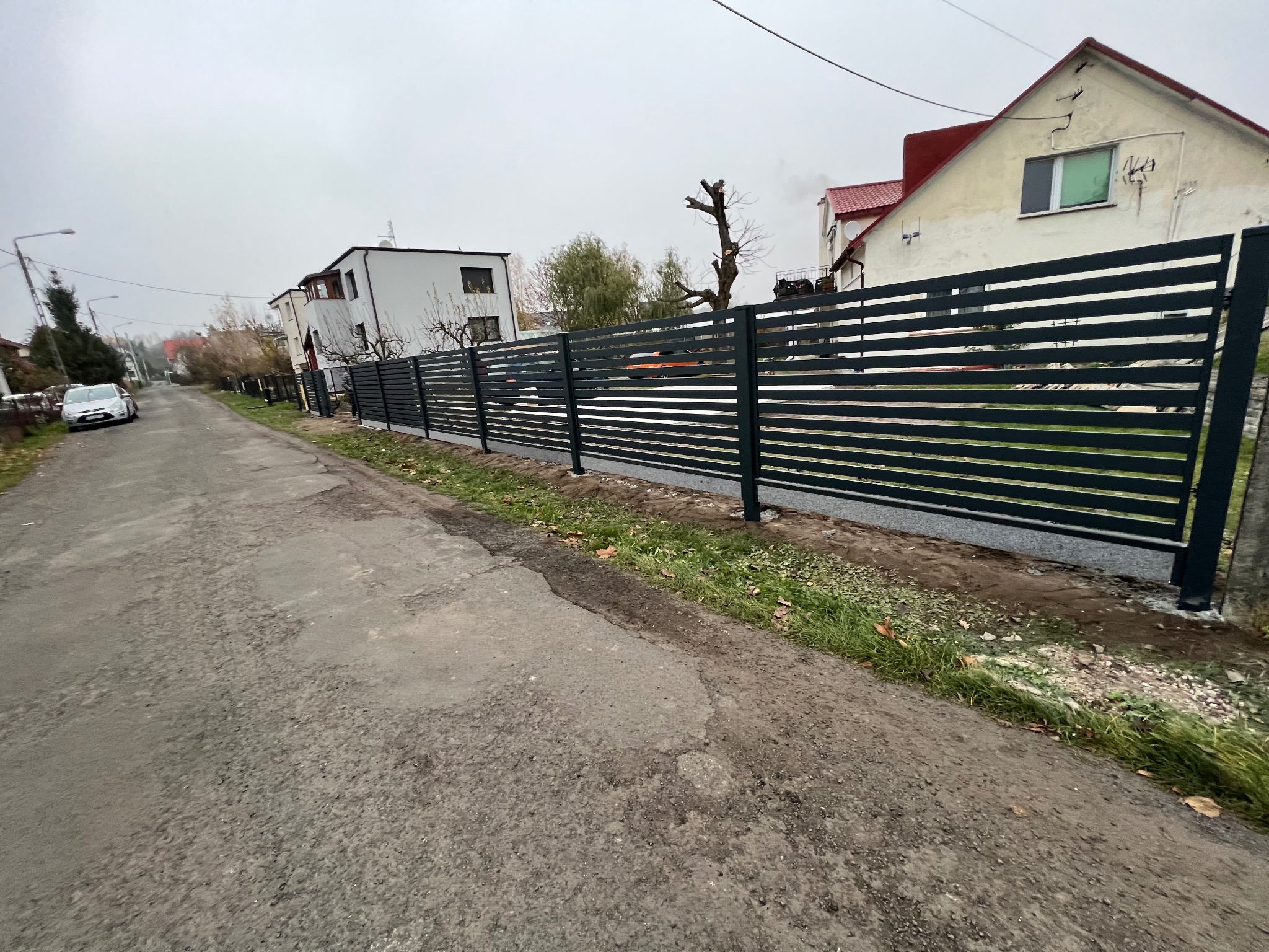 Nowoczesne, poziome, metalowe ogrodzenie panelowe w kolorze antracytowym, oddzielające posesję od drogi, z widocznymi domami w tle, sfotografowane w pochmurny dzień.
