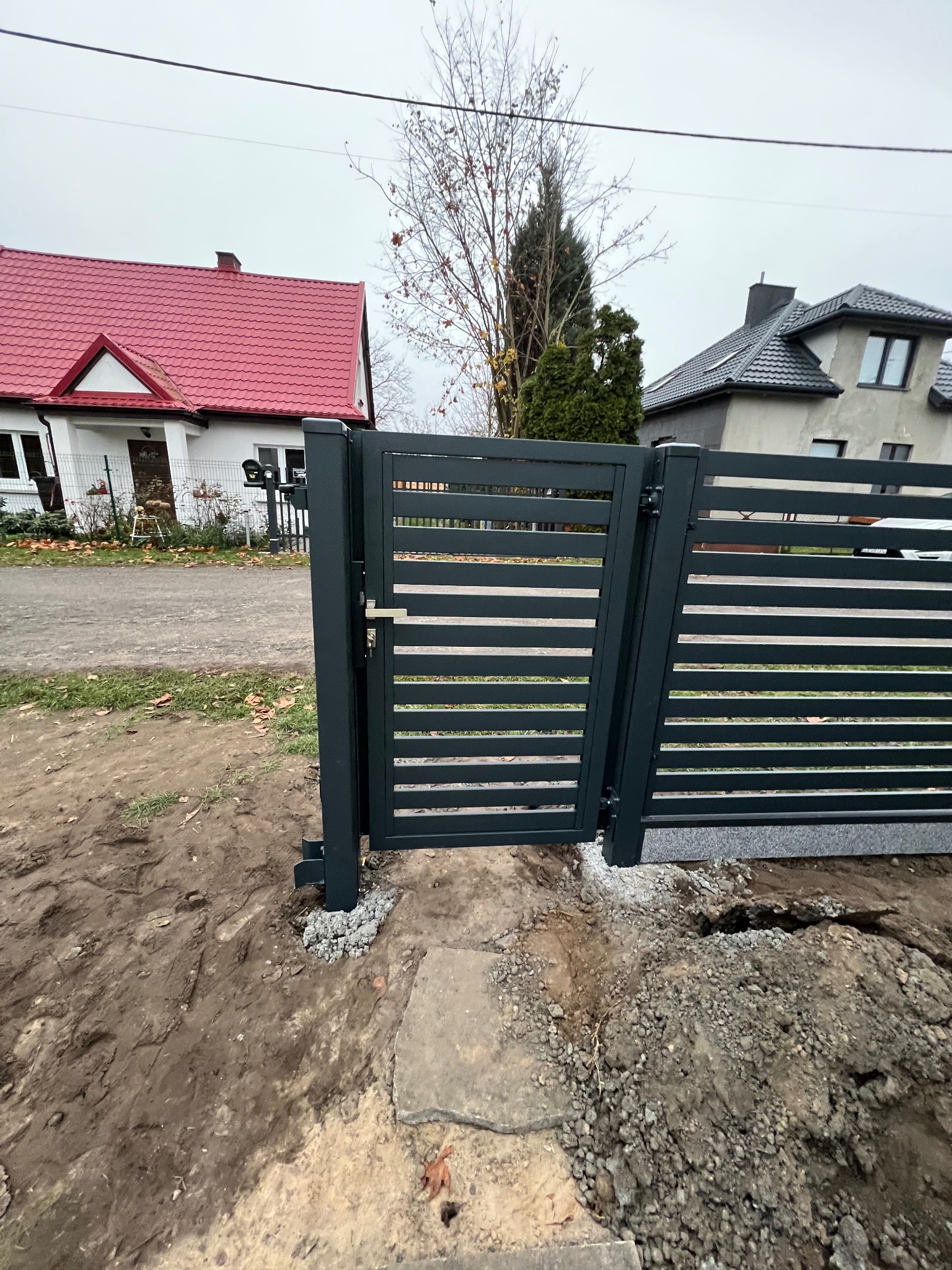 Nowoczesna, grafitowa brama wjazdowa z poziomymi szczeblami, częściowo otwarta, na tle dwóch domów jednorodzinnych w pochmurny dzień. Widoczne ślady prac ziemnych wokół bramy.