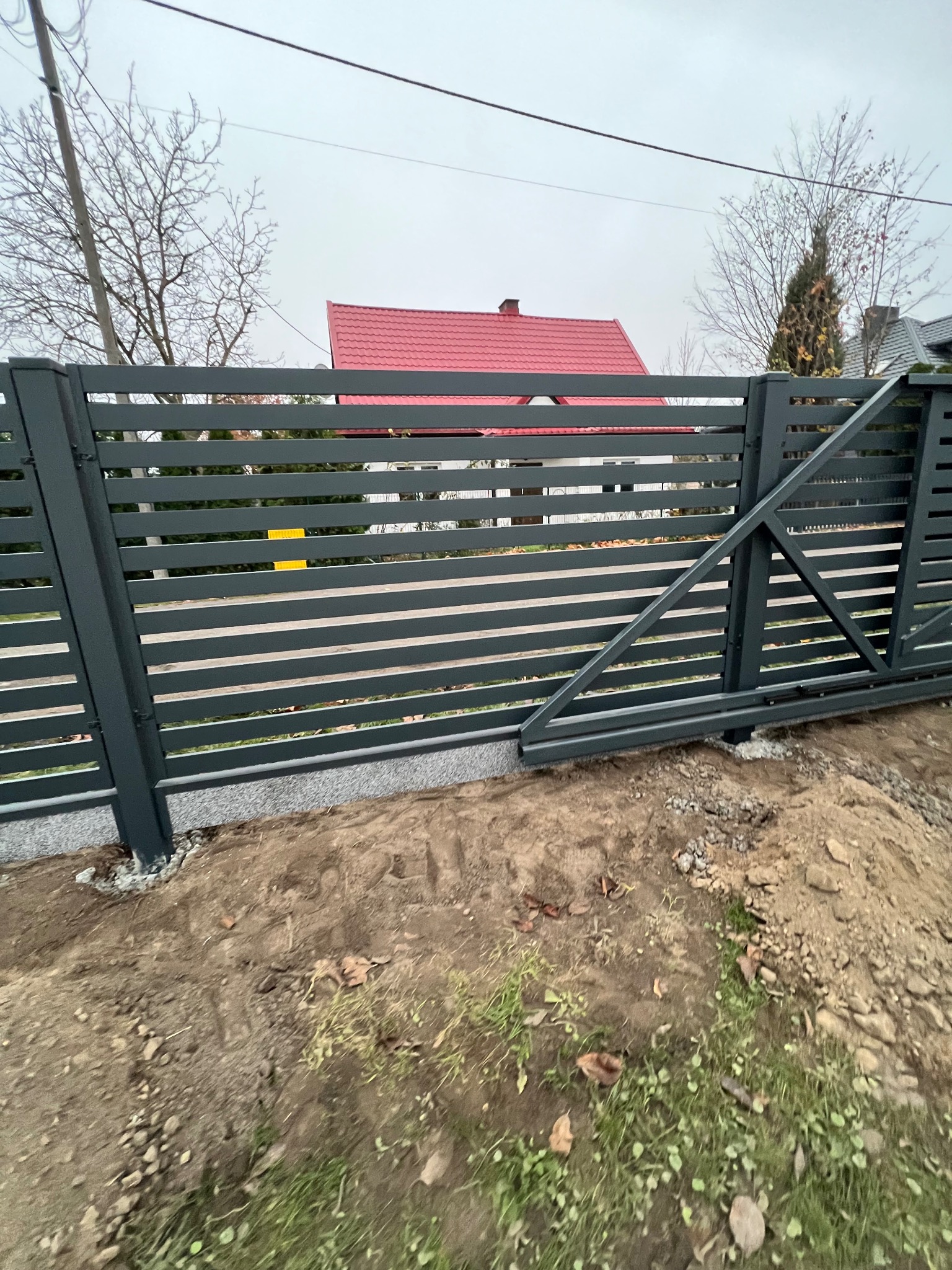Nowoczesna brama przesuwna z poziomych profili w kolorze grafitowym, widoczne fundamenty i przygotowany teren, w tle dom z czerwonym dachem.