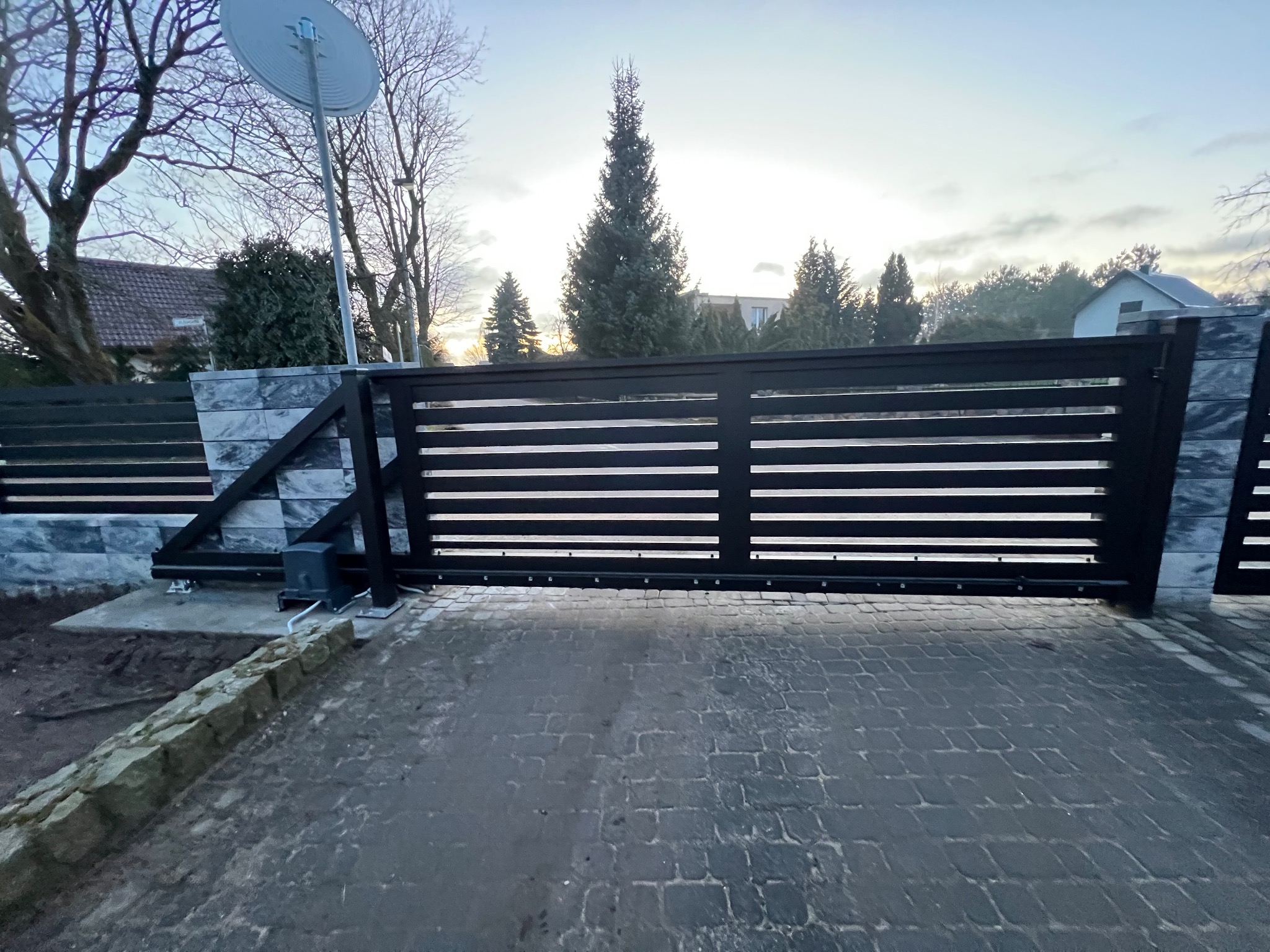Nowoczesna, przesuwna brama wjazdowa z poziomymi szczeblami, zamontowana na posesji z kostką brukową, widoczny napęd automatyczny i element ogrodzenia z imitacji kamienia.
