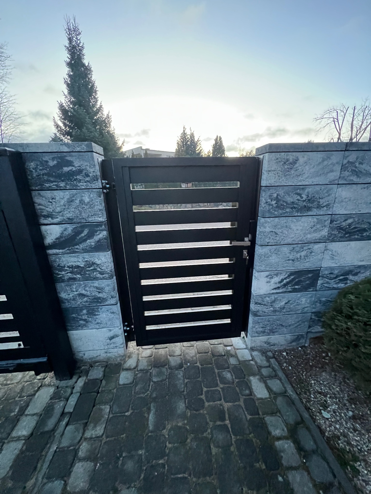 Nowoczesna, czarna furtka z poziomymi szczeblami, wkomponowana w ogrodzenie z szarej, łupanej kostki betonowej, z widocznym brukowanym chodnikiem i iglastym drzewem w tle.