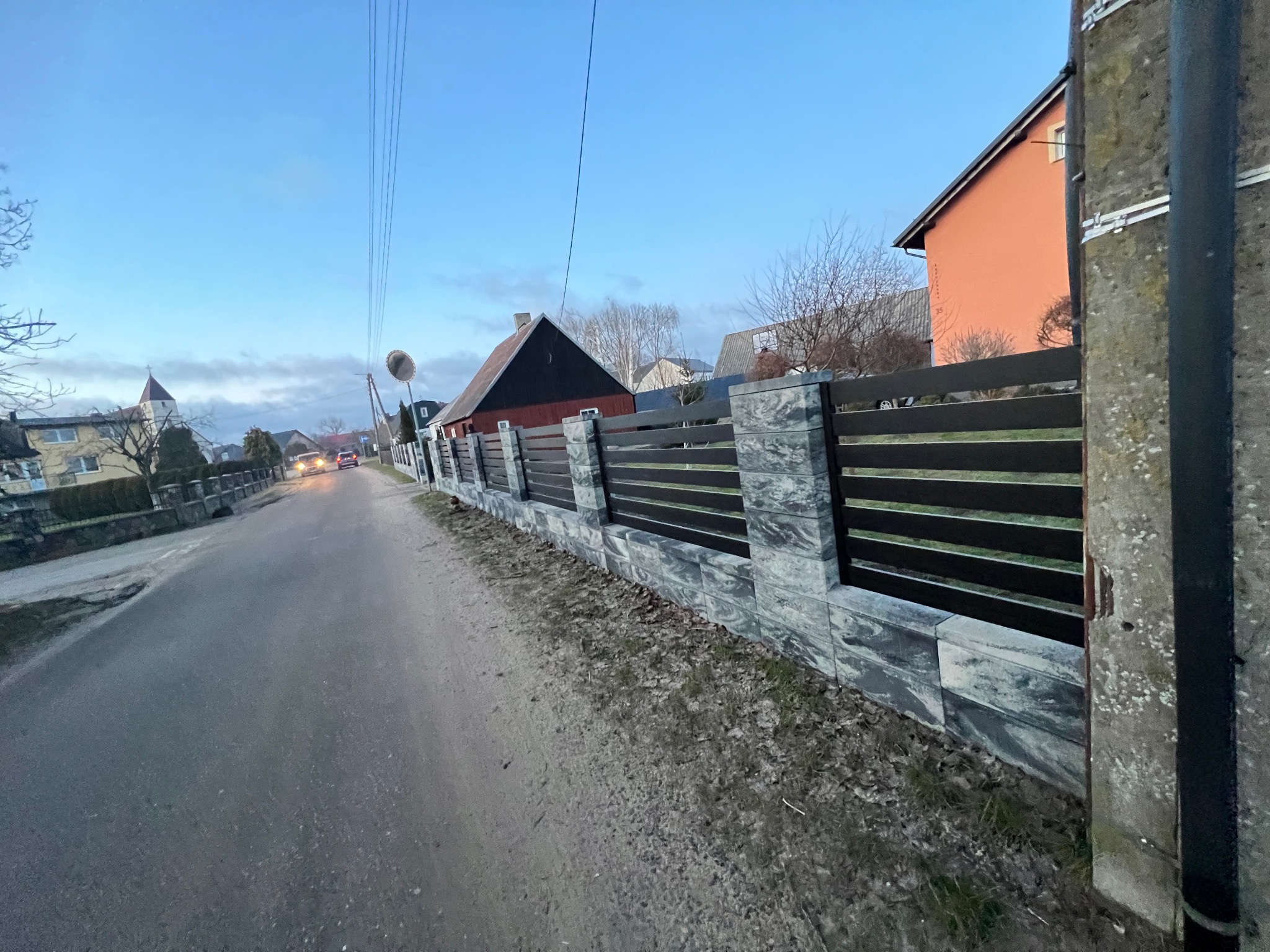 Nowoczesne ogrodzenie panelowe z elementami imitującymi kamień, widoczne z perspektywy ulicy w pochmurny dzień. W tle domy i przejeżdżający samochód.