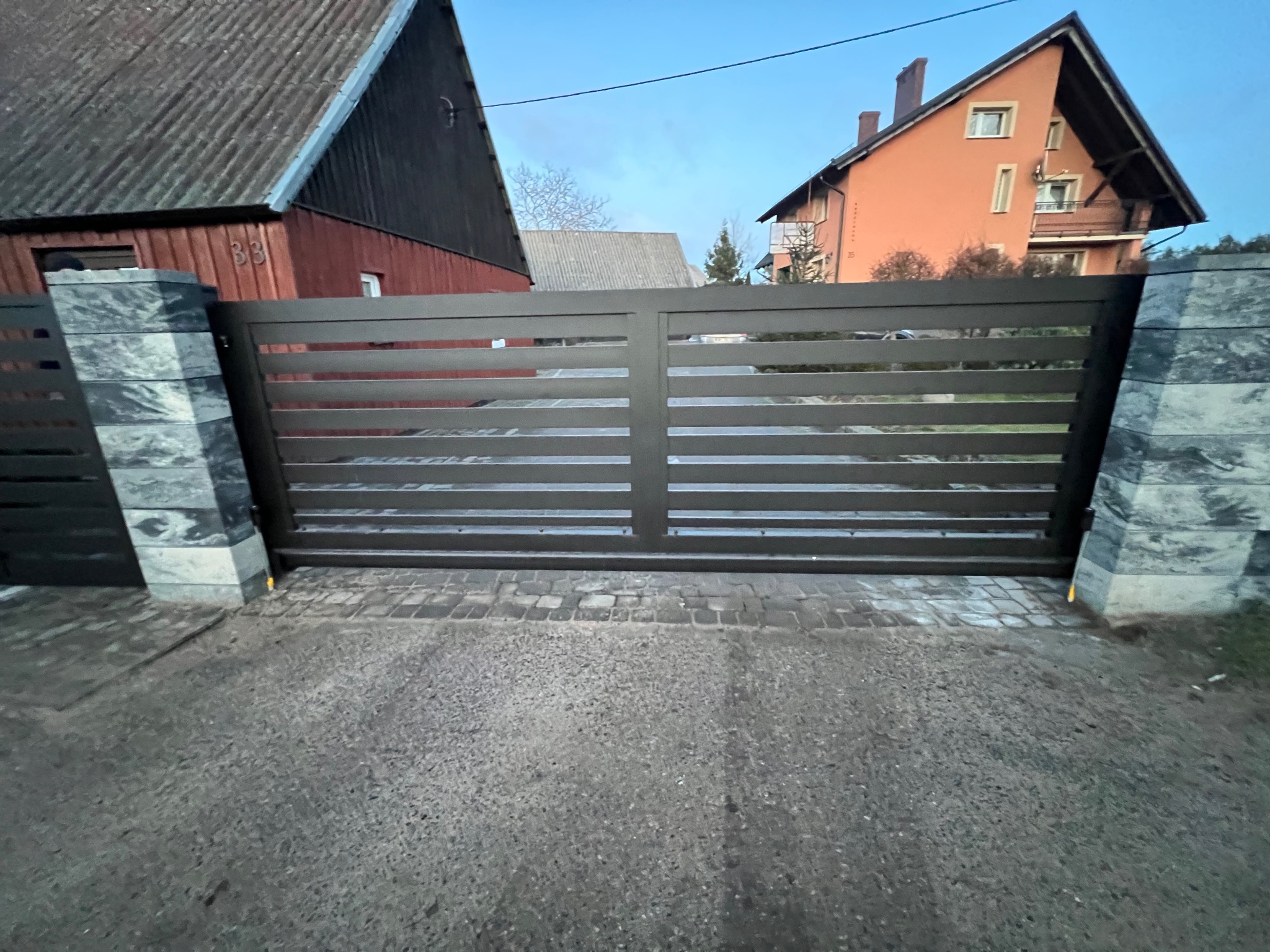 Nowoczesna, pozioma brama wjazdowa w kolorze antracytowym, osadzona na kamiennych słupkach z szarego kamienia łupanego, na tle zabudowań mieszkalnych i brukowanej nawierzchni.