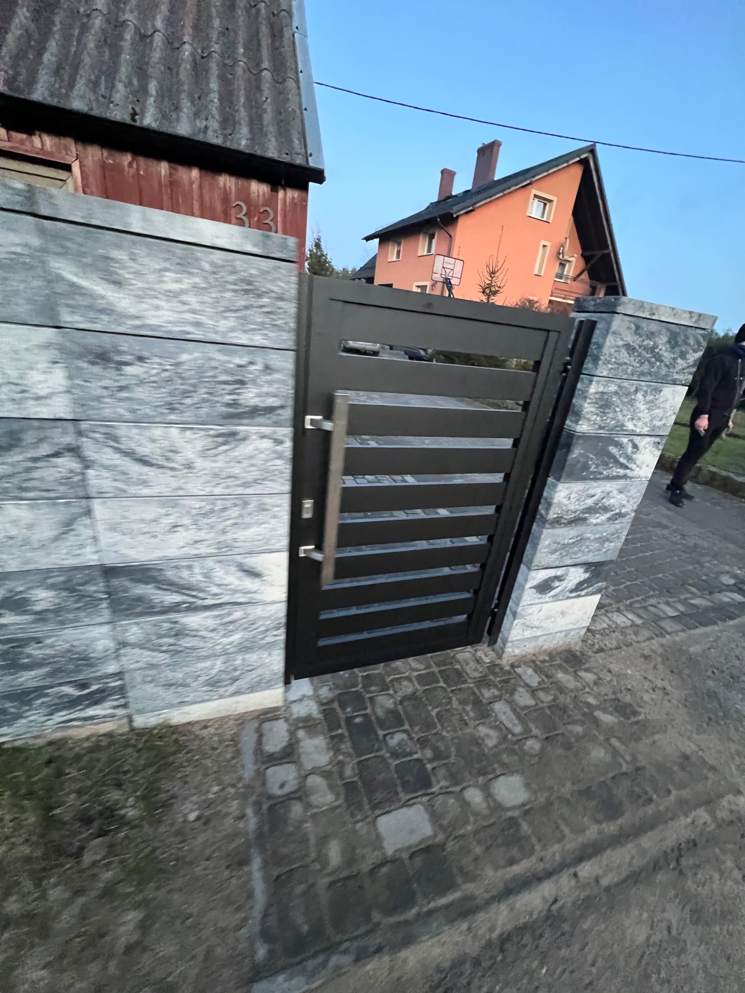 Nowoczesna czarna brama z poziomymi szczeblami, zamontowana w ogrodzeniu z szarych, kamiennych bloków. W tle widoczny dom z pomarańczową elewacją i fragment chodnika z kostki brukowej, osoba...