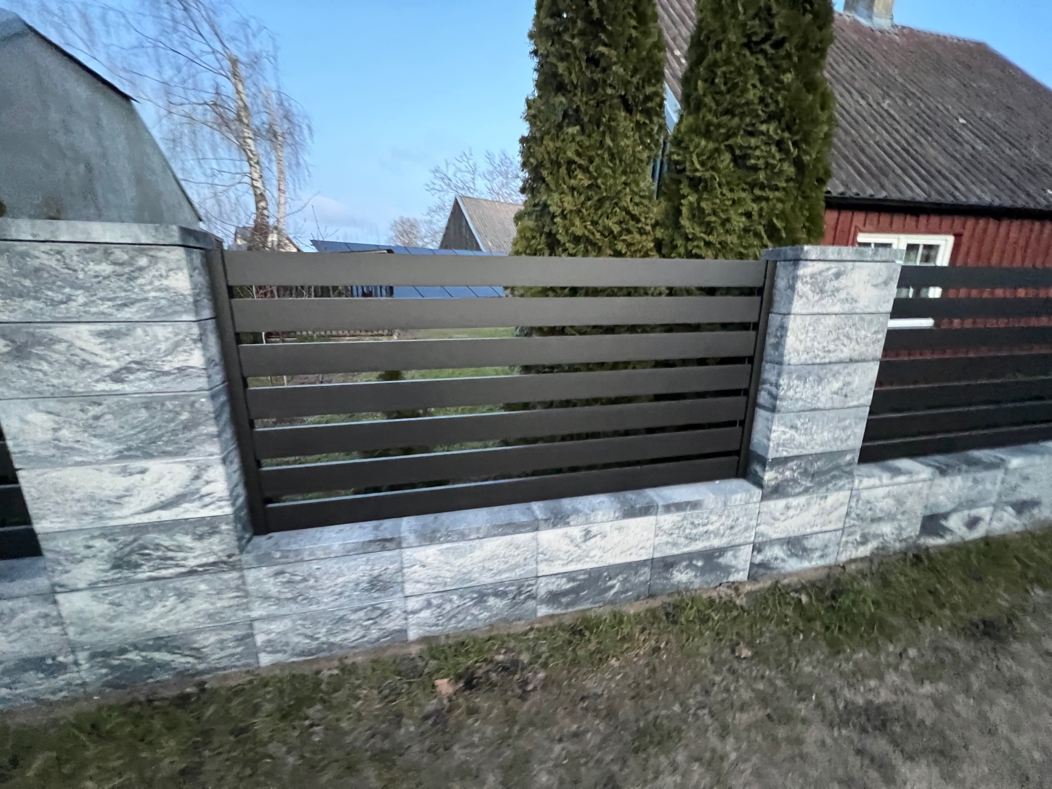 Nowoczesne ogrodzenie z poziomych, grafitowych paneli między słupkami z szarej, imitującej kamień kostki, na tle domów i drzew
