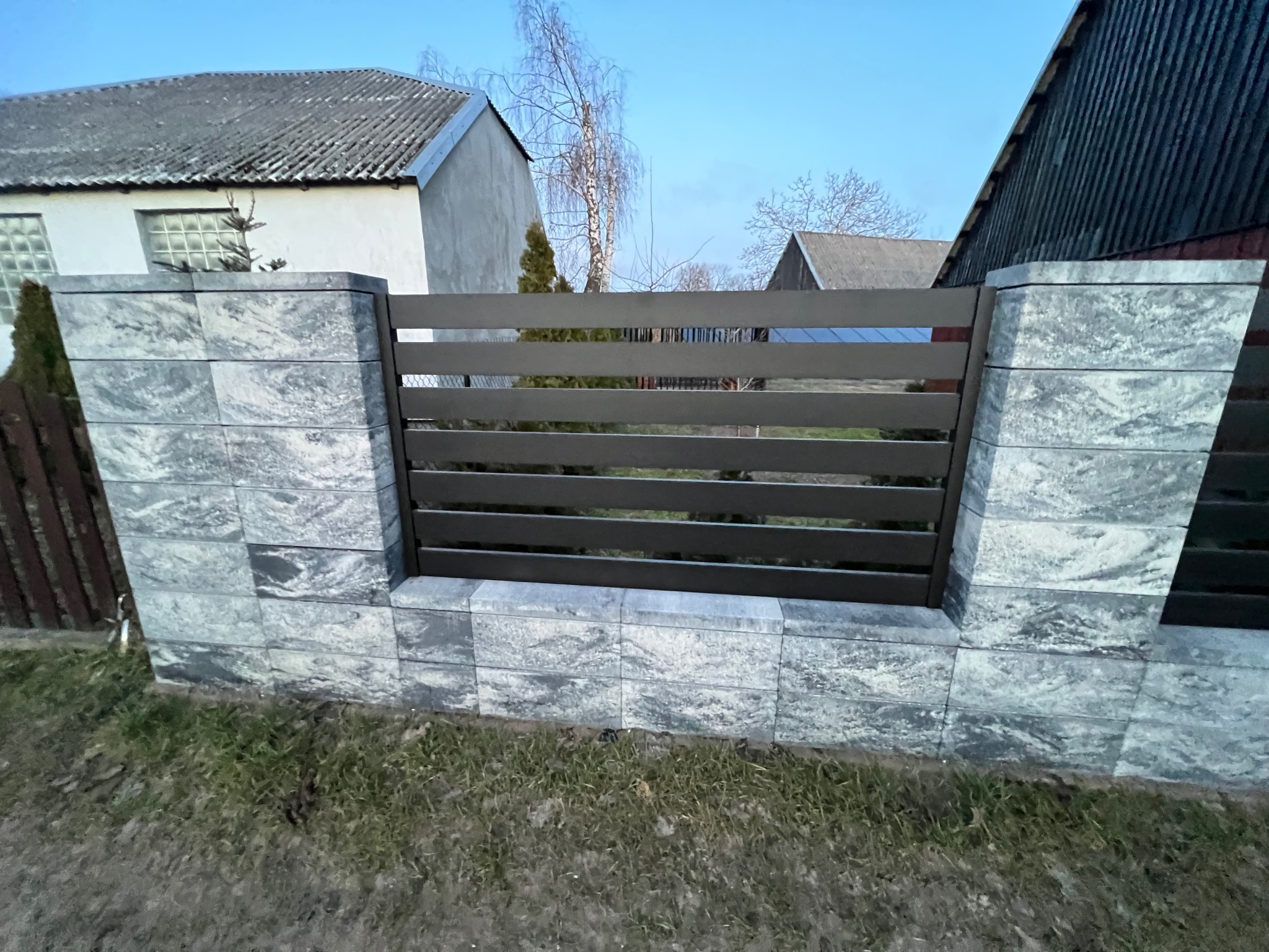 Nowoczesne ogrodzenie z szarych, betonowych bloczków i poziomych, metalowych przęseł na tle domów i błękitnego nieba.