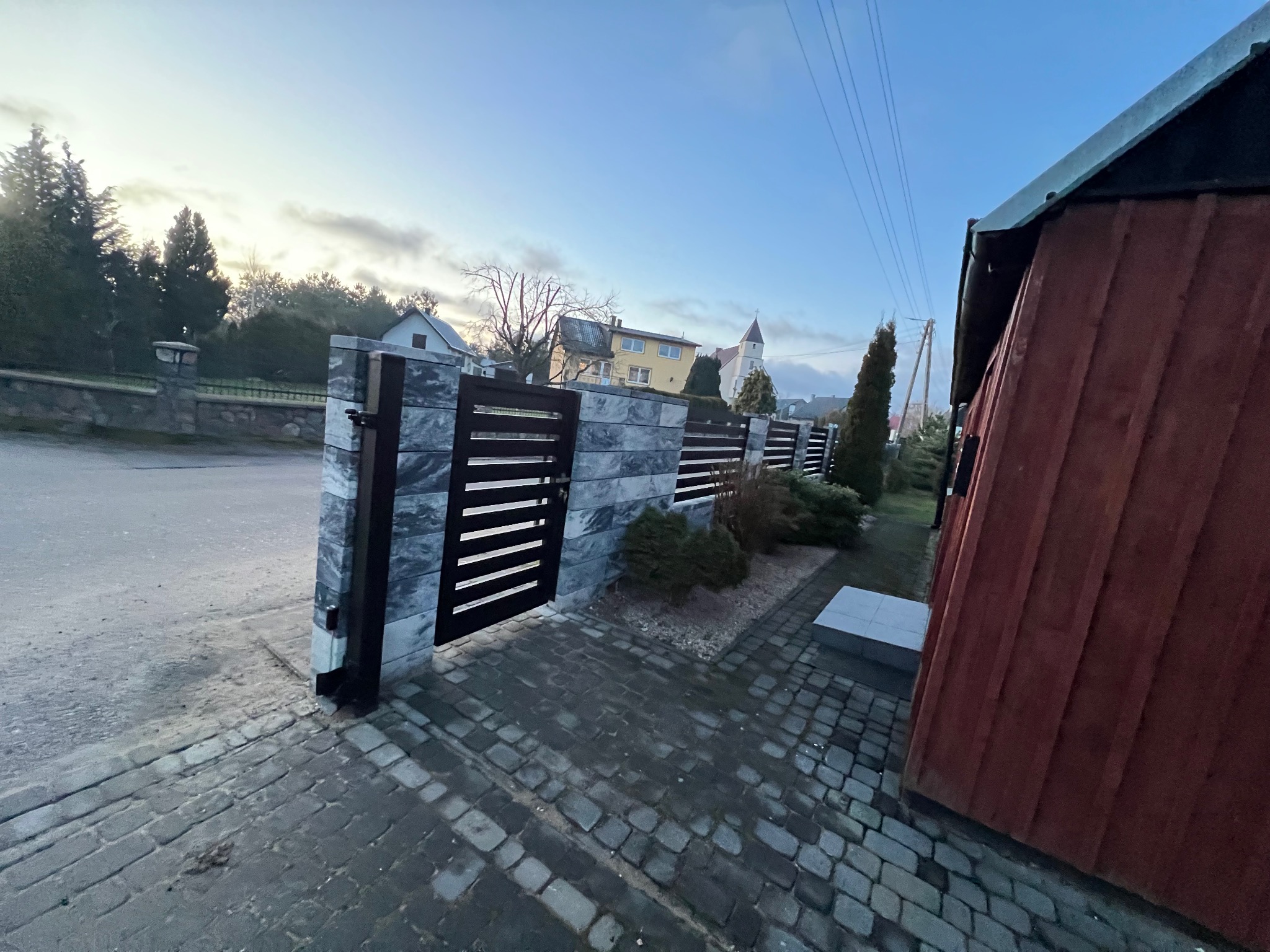 Nowoczesne ogrodzenie panelowe z elementami imitującymi kamień i poziomymi przęsłami w kolorze grafitowym, widoczne z perspektywy chodnika wyłożonego kostką brukową, w tle budynki mieszkalne...