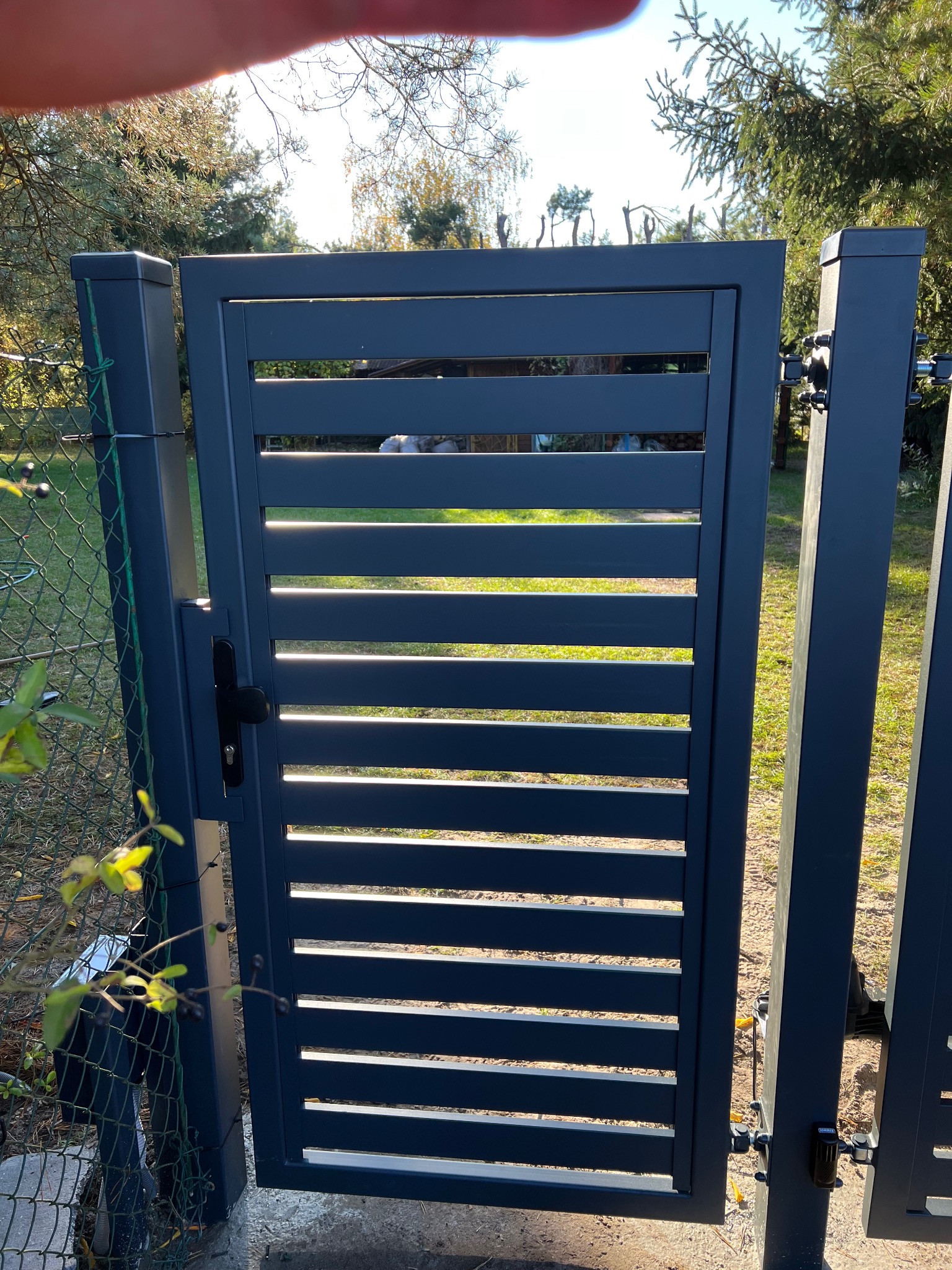 Nowoczesna, antracytowa brama lamelowa z czarną klamką, zamontowana w ogrodzeniu z siatki, z widoczną automatyką i zaroślami w tle.