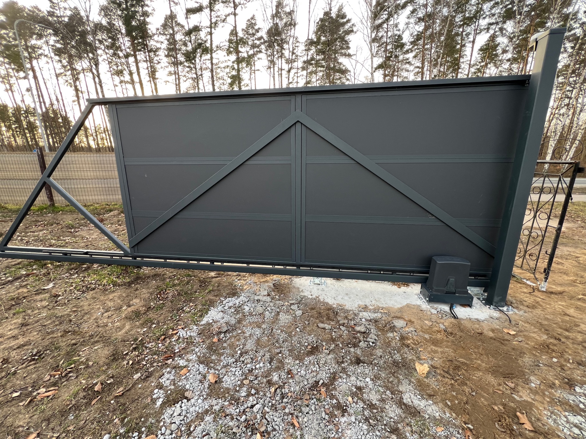 Przesuwna brama wjazdowa w kolorze grafitowym z widocznym napędem automatycznym, zamontowana na betonowym fundamencie, w tle drzewa.
