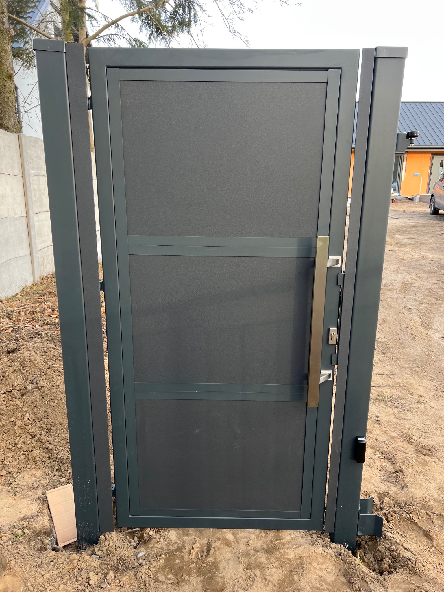 Nowoczesna, szara furtka ogrodzeniowa z metalową ramą i długim, pionowym uchwytem, zamontowana w ziemi, widoczny czytnik zbliżeniowy i elementy montażowe w ziemi.