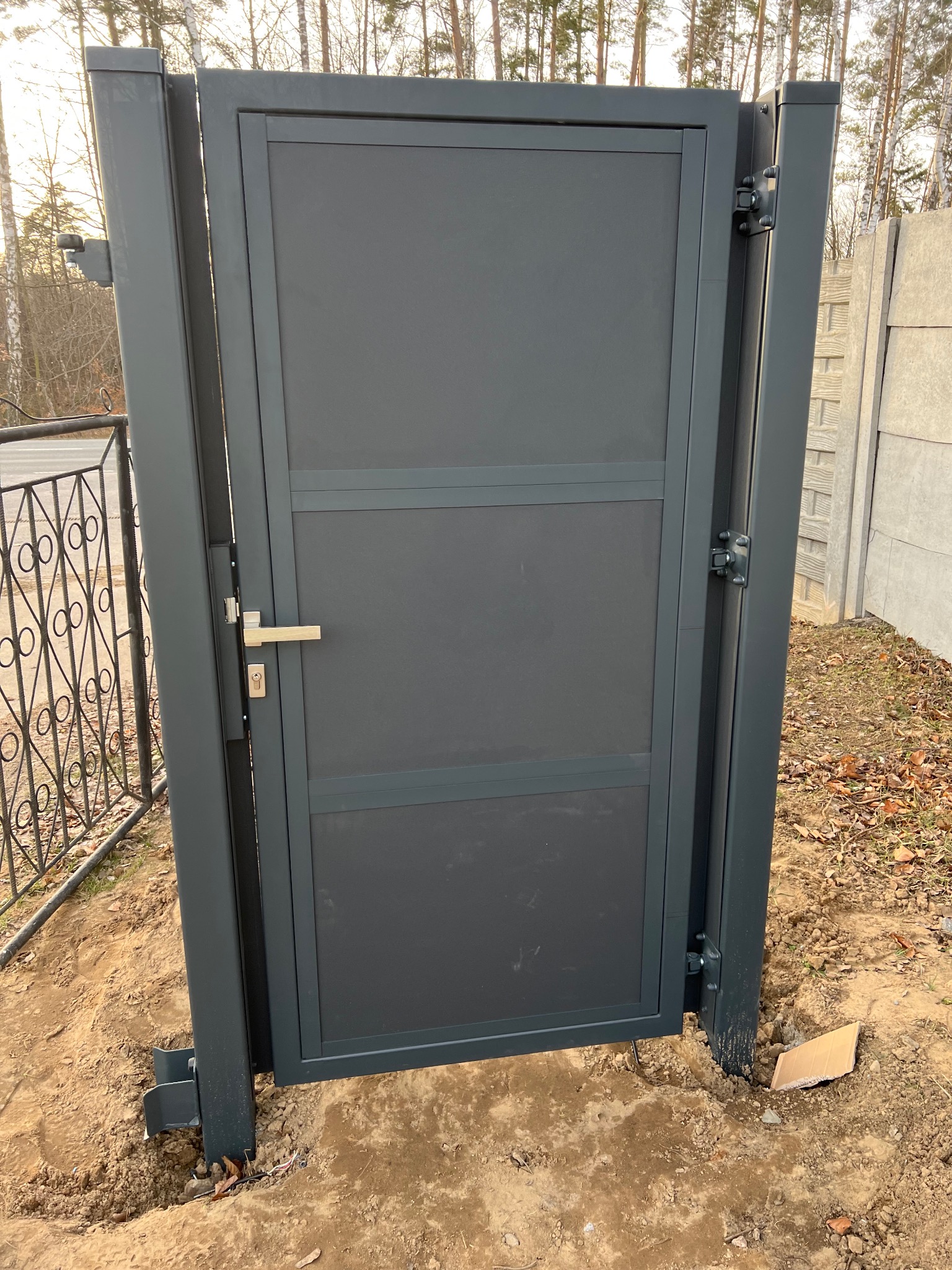 Grafitowa furtka z metalową ramą i srebrną klamką, osadzona w ziemi, widoczne słupki, zawiasy i fragment ogrodzenia panelowego w tle.