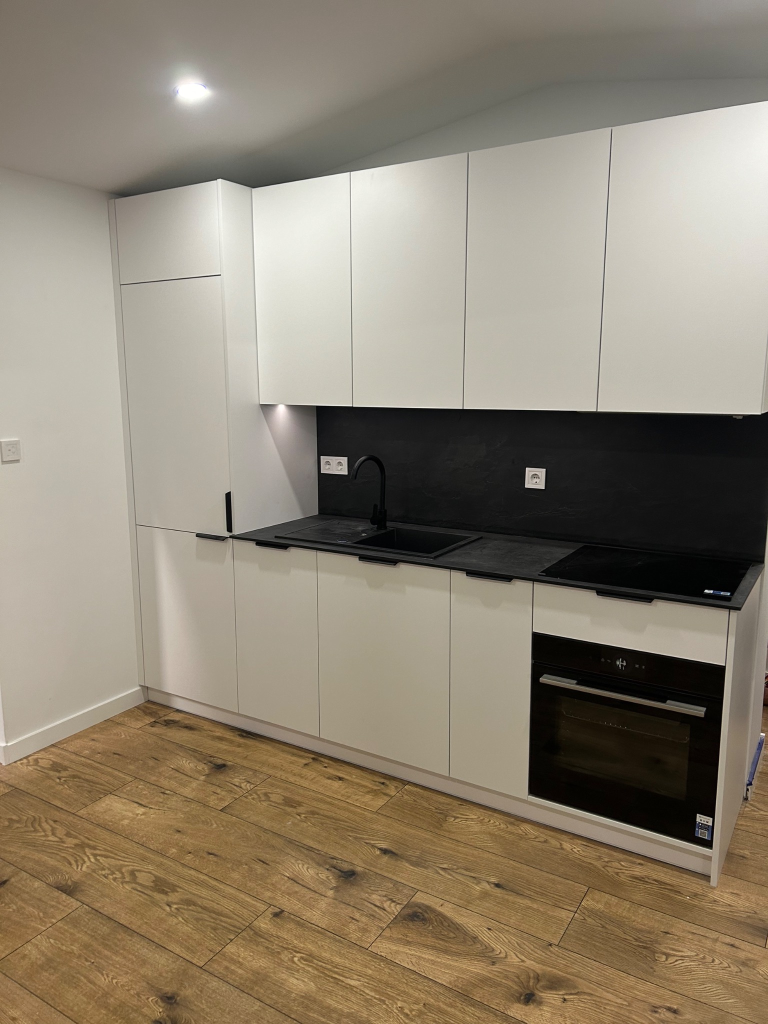 Nowoczesna, minimalistyczna kuchnia z białymi frontami i czarnymi akcentami: bateria, panel nad blatem, płyta indukcyjna i piekarnik. Drewniana podłoga dodaje ciepła.