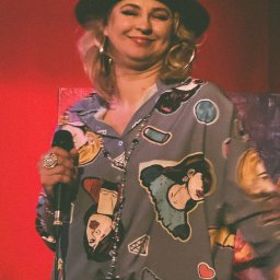 Łukasz Zajączkowski Fotografia - Uśmiechnięta kobieta w czerwonym kapeluszu i wzorzystej koszuli, śpiewająca do mikrofonu z dużym pierścieniem, na czerwonym tle.