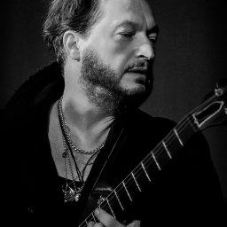 Łukasz Zajączkowski Fotografia - Czarno-białe portretowe ujęcie muzyka z brodą, grającego na gitarze akustycznej, z widocznymi pierścieniami i łańcuchami na szyi.