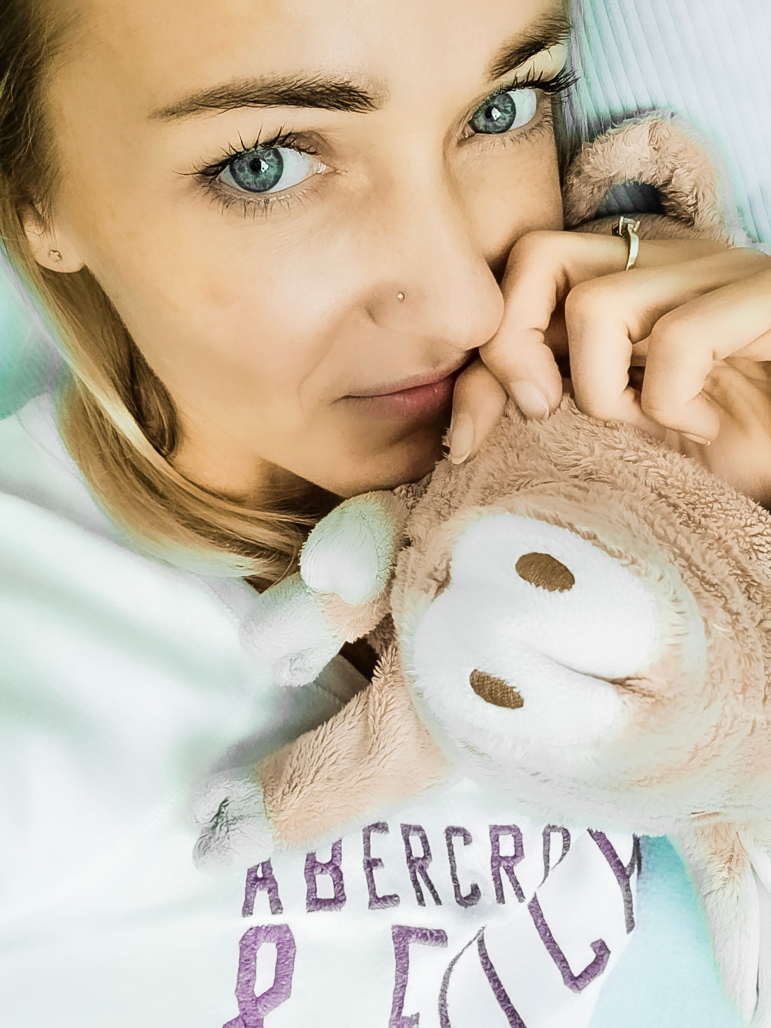 Portret kobiety z niebieskimi oczami, z delikatnym makijażem, trzymającej pluszową zabawkę, pierścionek na palcu, bluzka z napisem.