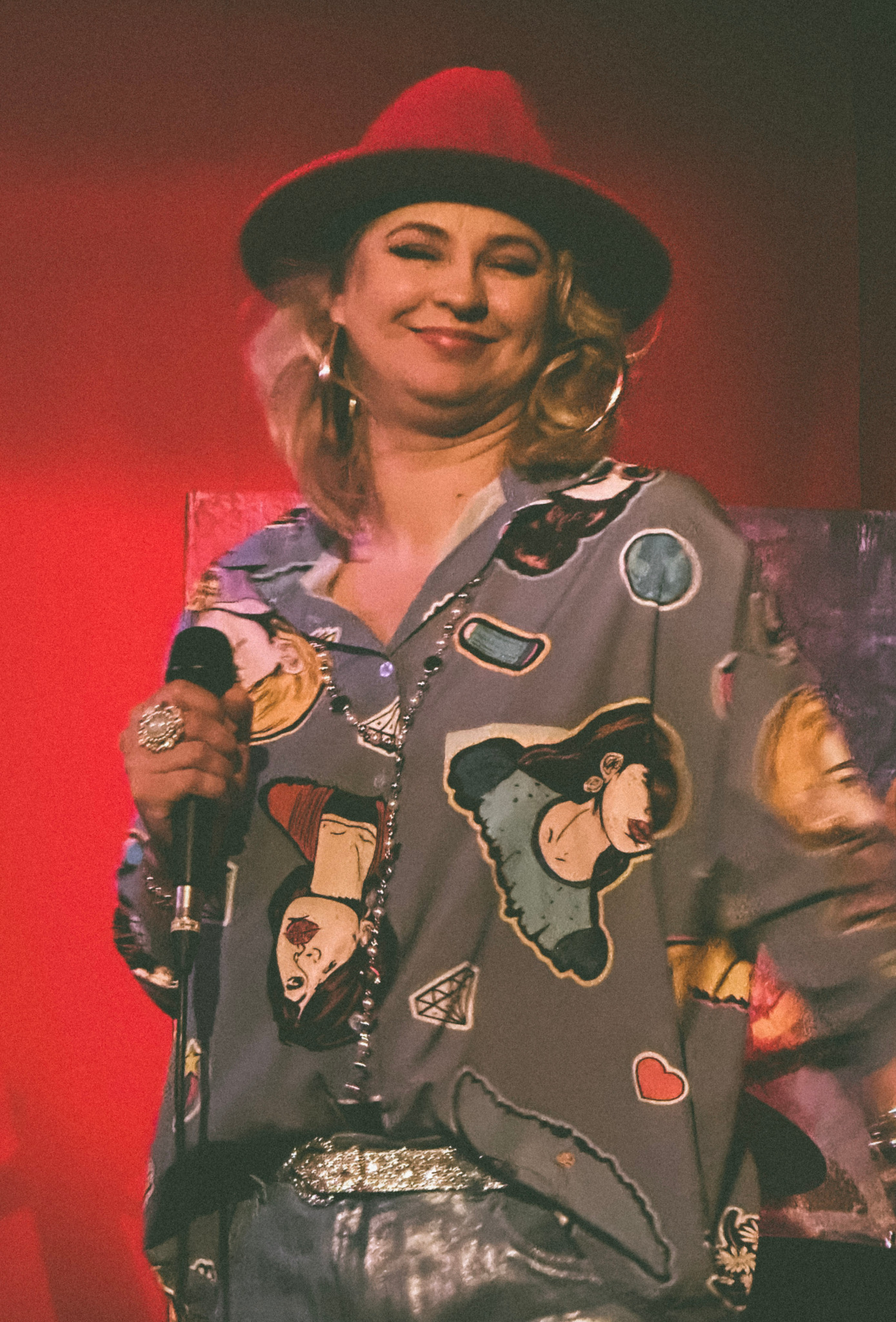 Uśmiechnięta kobieta w czerwonym kapeluszu i wzorzystej koszuli, śpiewająca do mikrofonu z dużym pierścieniem, na czerwonym tle.