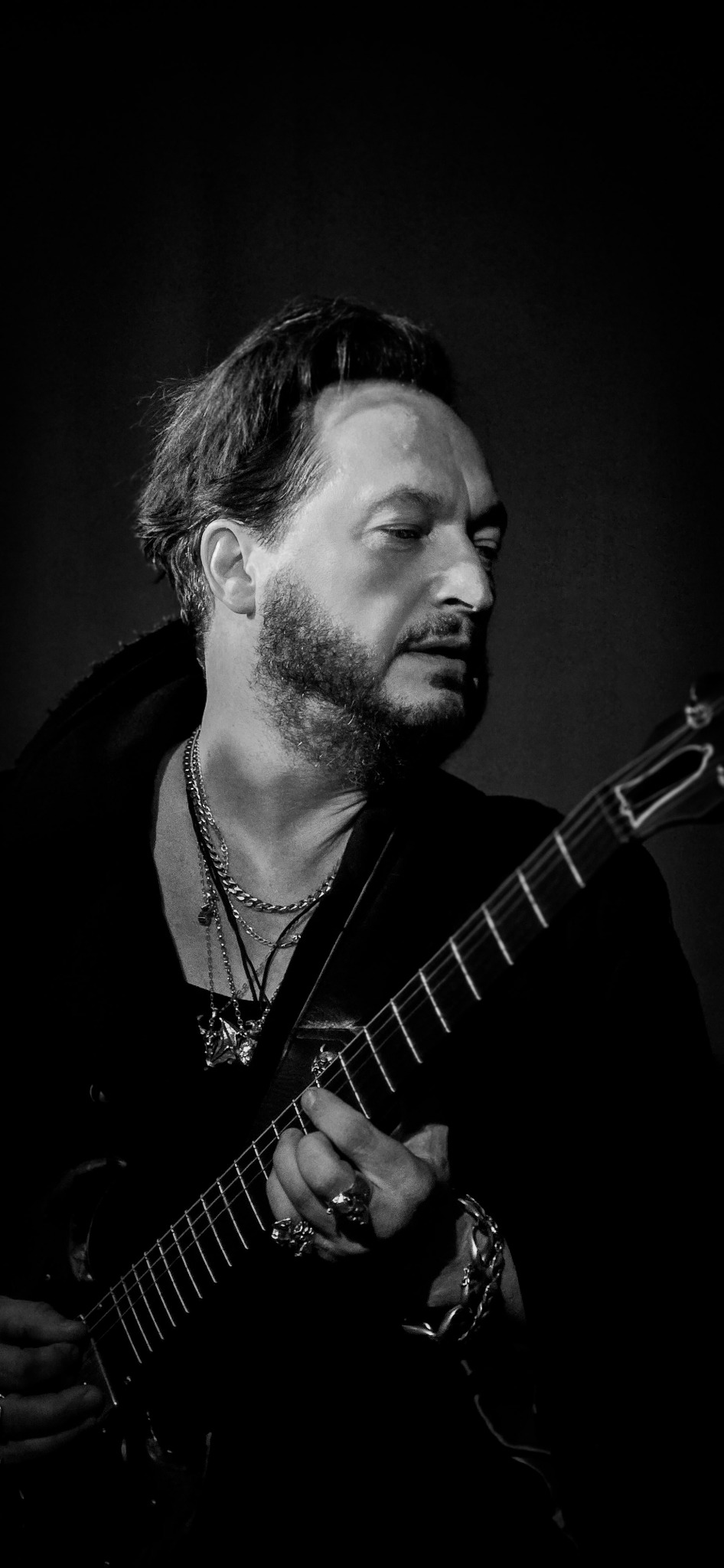 Czarno-białe portretowe ujęcie muzyka z brodą, grającego na gitarze akustycznej, z widocznymi pierścieniami i łańcuchami na szyi.
