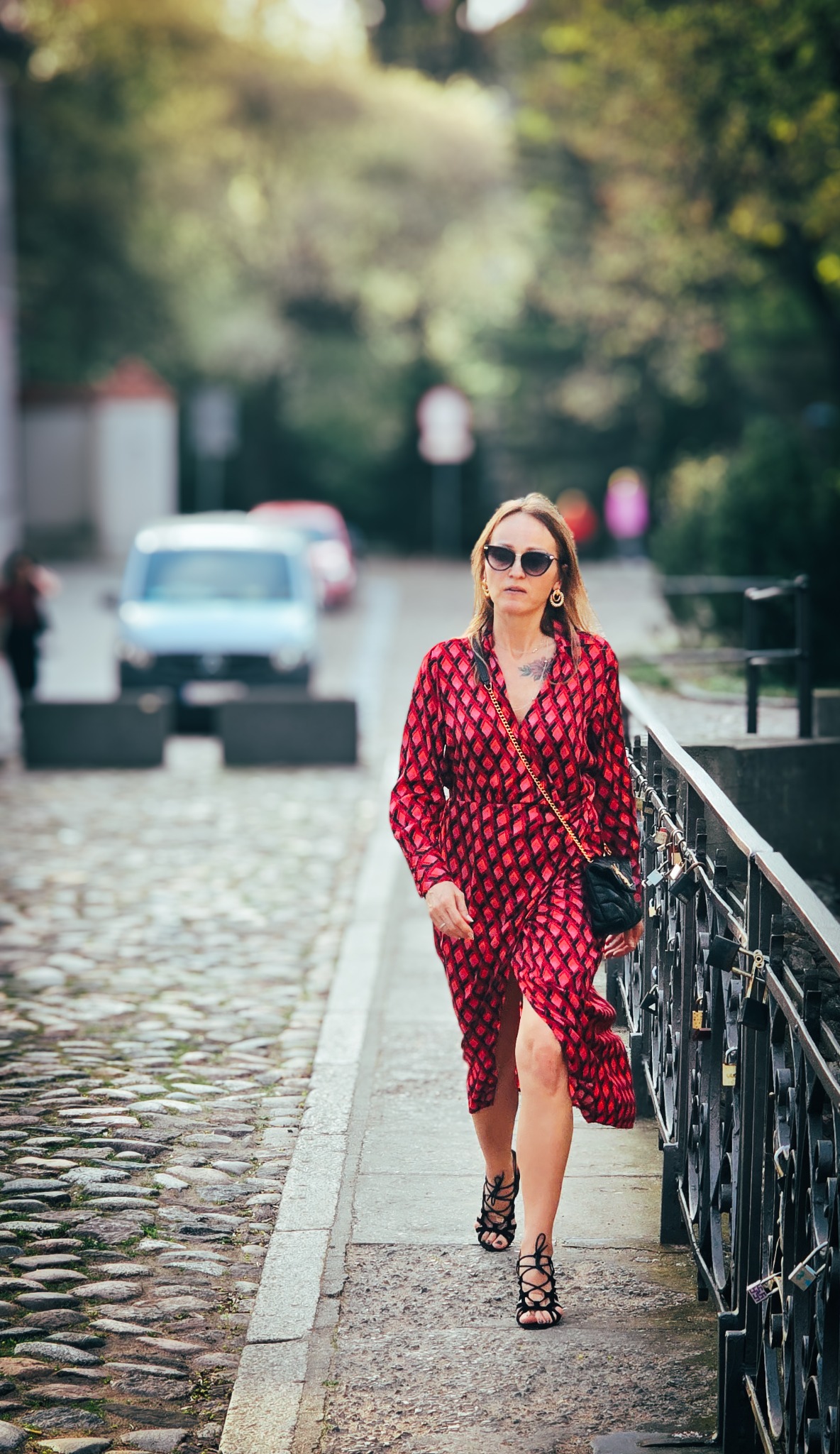 Kobieta w czerwonej wzorzystej sukience i czarnych sandałach na obcasie spaceruje po chodniku wzdłuż metalowej barierki z kłódkami, z rozmyciem w tle.