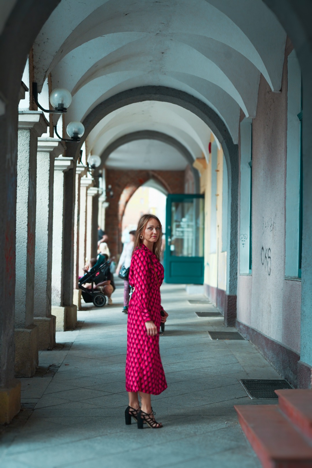 Kobieta w wzorzystej sukience w różowo-czerwonej kolorystyce, stojąca w podcieniach z łukowym sklepieniem, spoglądająca w obiektyw