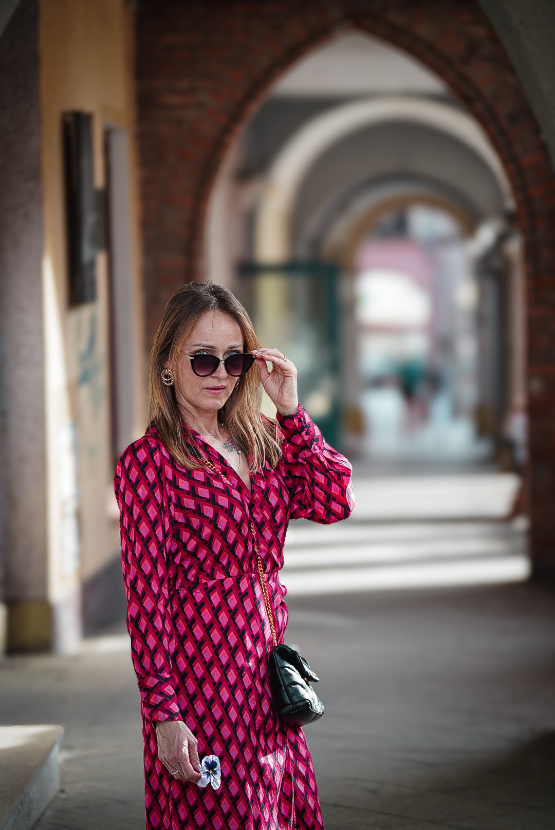 Kobieta w okularach przeciwsłonecznych, w różowo-czarnej sukience w romby, pozuje w przejściu z ceglanymi łukami, trzymając kwiatek w dłoni.