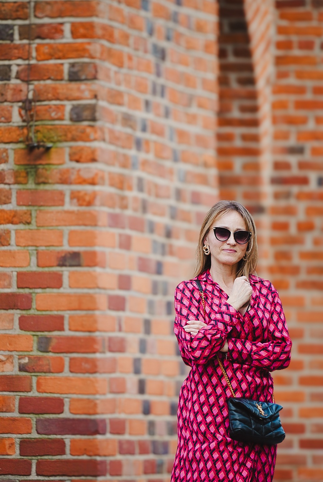 Kobieta w okularach przeciwsłonecznych pozuje na tle ceglanej ściany w Olsztynie, ubrana w różową sukienkę w romby i czarną torebkę na złotym łańcuszku.