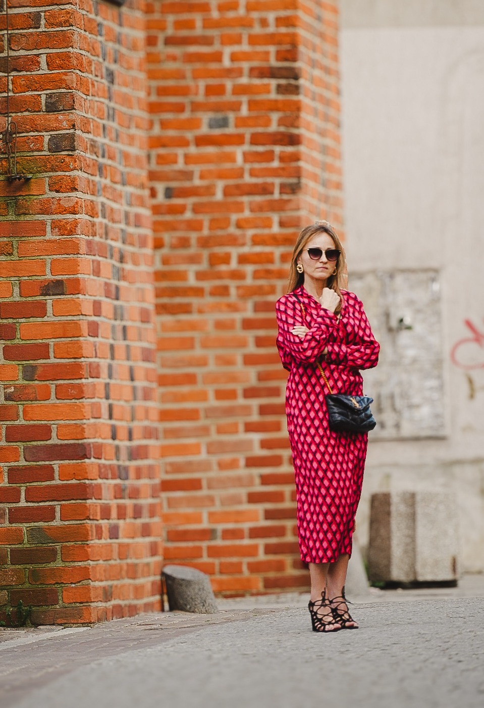 Kobieta w różowej sukience w romby, czarnych sandałach na obcasie i okularach przeciwsłonecznych pozuje na tle ceglanej ściany i jasnej elewacji z graffiti.
