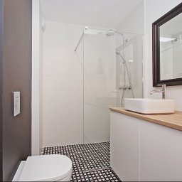 Dessein Magdalena Pawlak - Nowoczesna łazienka z kabiną prysznicową ze szklaną ścianką, białą umywalką nablatową na szafce z drewnianym blatem, dużym lustrem w ciemnej ramie i czarno-białą mozaiką na podłodze.