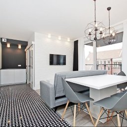 Dessein Magdalena Pawlak - Jasne wnętrze mieszkania z aneksem kuchennym, białymi meblami, ścianą z białej cegły, szarym narożnikiem i stołem z krzesłami. Nad stołem wiszą dwie nowoczesne, druciane lampy. Na podłodze...