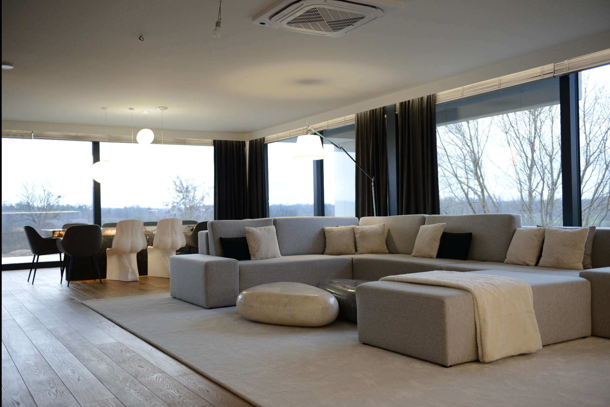 Jasne, przestronne wnętrze salonu z modułową sofą w odcieniach szarości, ozdobnymi poduszkami, kamiennym stolikiem kawowym i widokiem na zewnątrz przez duże okna z ciemnymi zasłonami.
