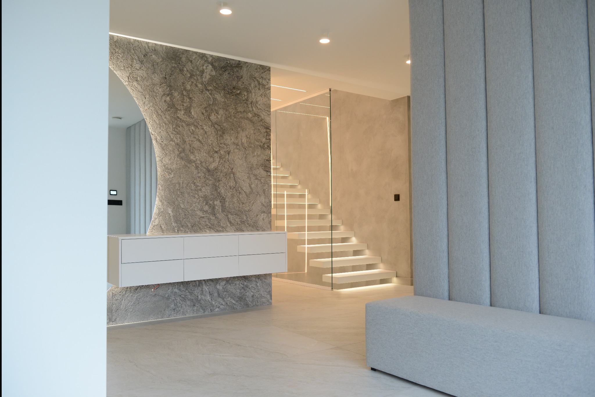 Nowoczesne wnętrze mieszkania z podświetlanymi schodami, ścianą z efektem kamienia i minimalistyczną białą komodą wiszącą, uzupełnione szarą tapicerowaną ścianą i siedziskiem.