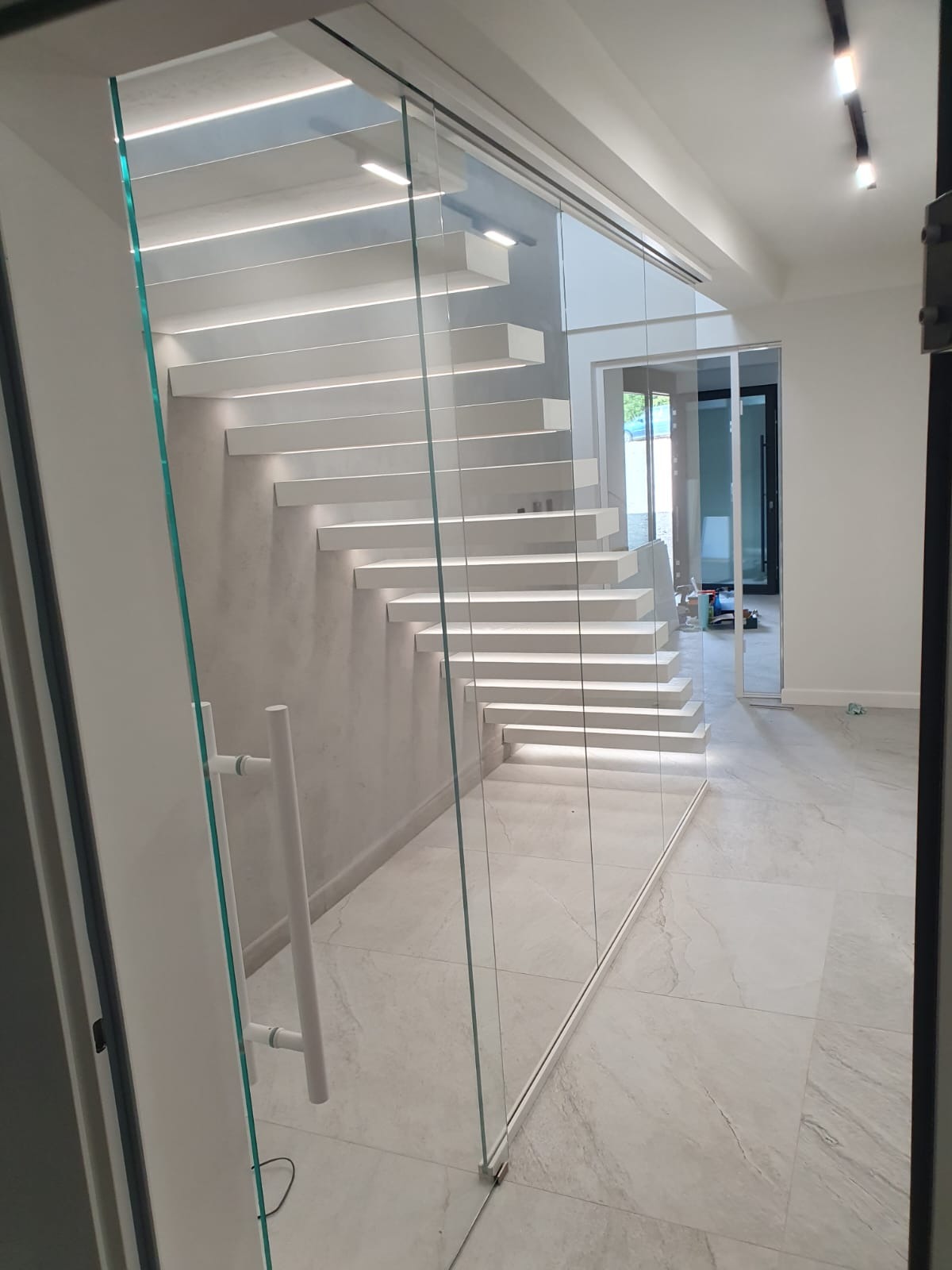 Nowoczesne schody dywanowe, białe stopnie podświetlone od spodu, szklana balustrada z minimalistycznymi uchwytami, marmurowa posadzka, widok z boku przez otwarte drzwi.