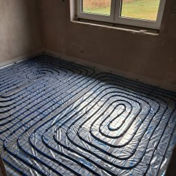 TWÓJ HYDRAULIK - Niebieskie rury ogrzewania podłogowego ułożone na folii z nadrukiem 'Kinstale' w surowym wnętrzu z oknem, widok z góry.