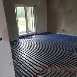 TWÓJ HYDRAULIK - Niebieskie rury ogrzewania podłogowego ułożone na folii w surowym wnętrzu z betonowymi ścianami i wylewką, widoczne okno balkonowe.