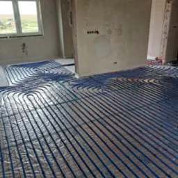 Niebieskie rury ogrzewania podłogowego ułożone na izolacji z siatką w surowym wnętrzu, widok z góry na dużą powierzchnię, okno w tle.