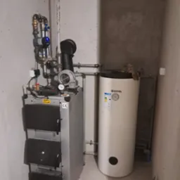 Kocioł na paliwo stałe z podajnikiem i zasobnikiem ciepłej wody użytkowej w kotłowni, widoczne rury instalacji grzewczej, manometr, wentylator i osprzęt.