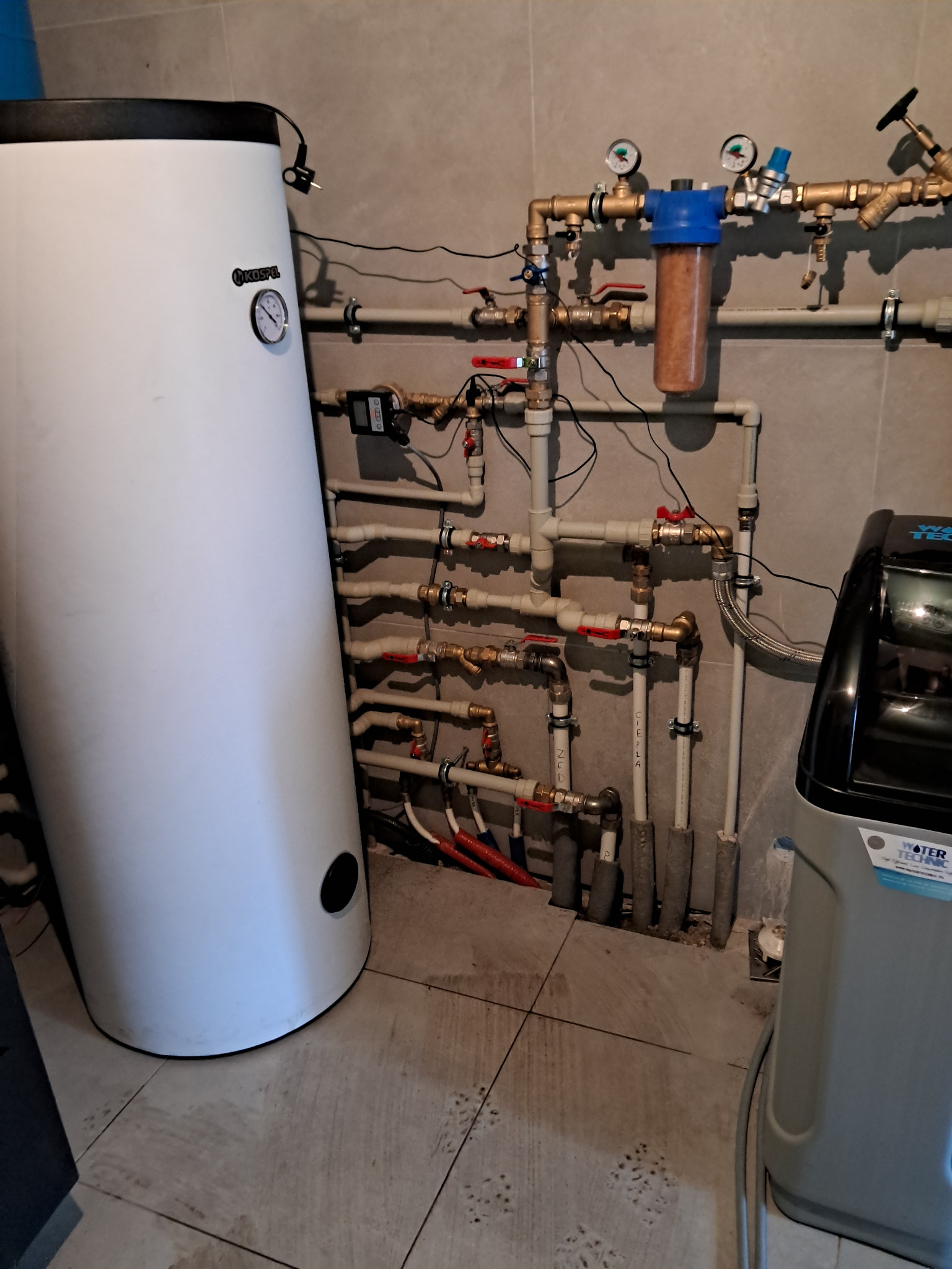 Biały bojler Kospel z manometrem, obok filtry wody i rozbudowana instalacja hydrauliczna z rurami PP, zaworami kulowymi i izolacją na rurach pionowych, na tle szarej ściany z płytek.