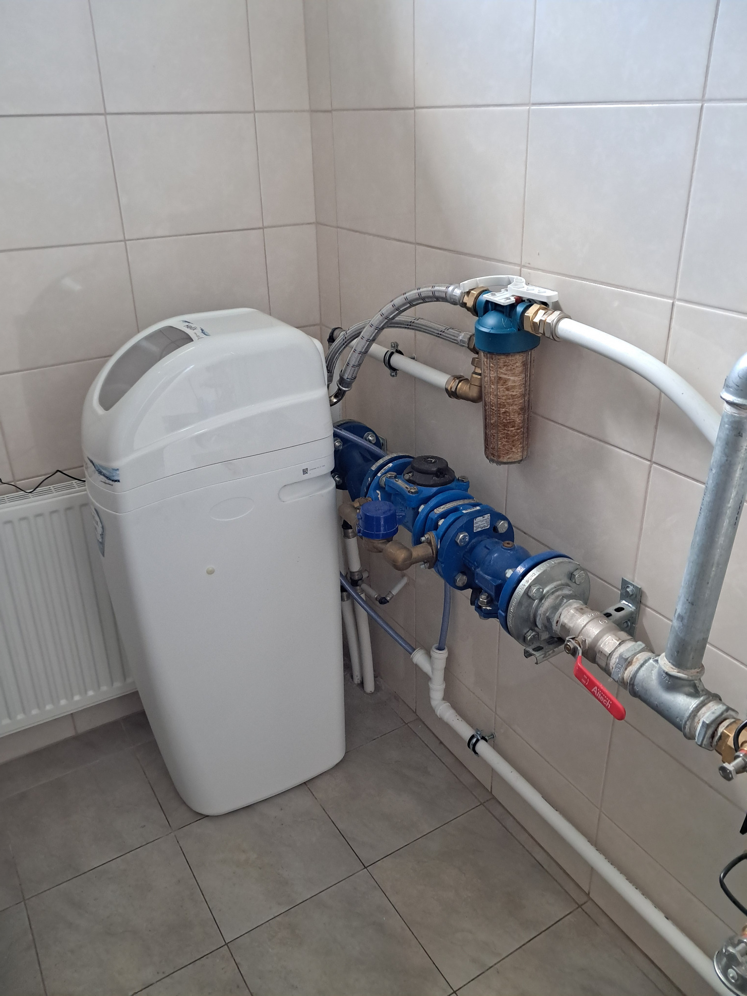 Biały zmiękczacz wody z filtrem mechanicznym i niebieską armaturą na tle ceramicznych płytek ściennych i podłogowych.