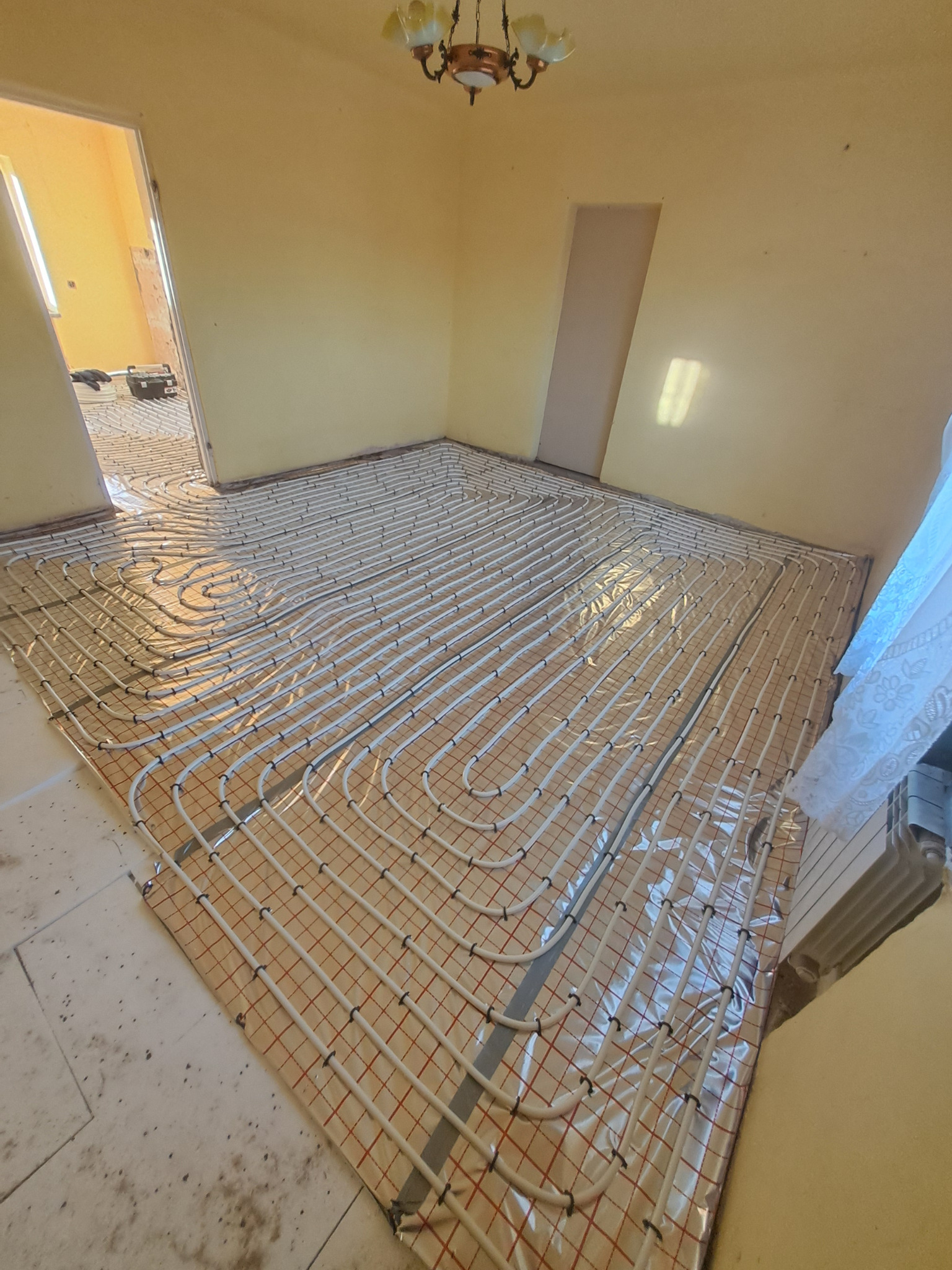 Rozłożone rury ogrzewania podłogowego na folii z siatką w jasnym pomieszczeniu, widoczny grzejnik i fragment instalacji w sąsiednim pokoju.
