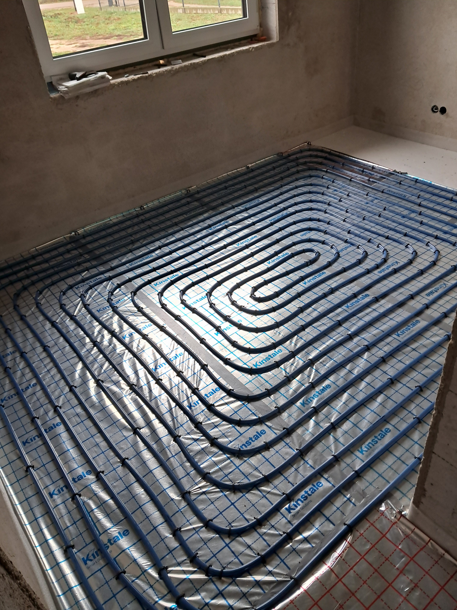Niedogrzane pomieszczenie w trakcie budowy z widocznymi rurami ogrzewania podłogowego marki Kinstale, ułożonymi spiralnie na izolacji