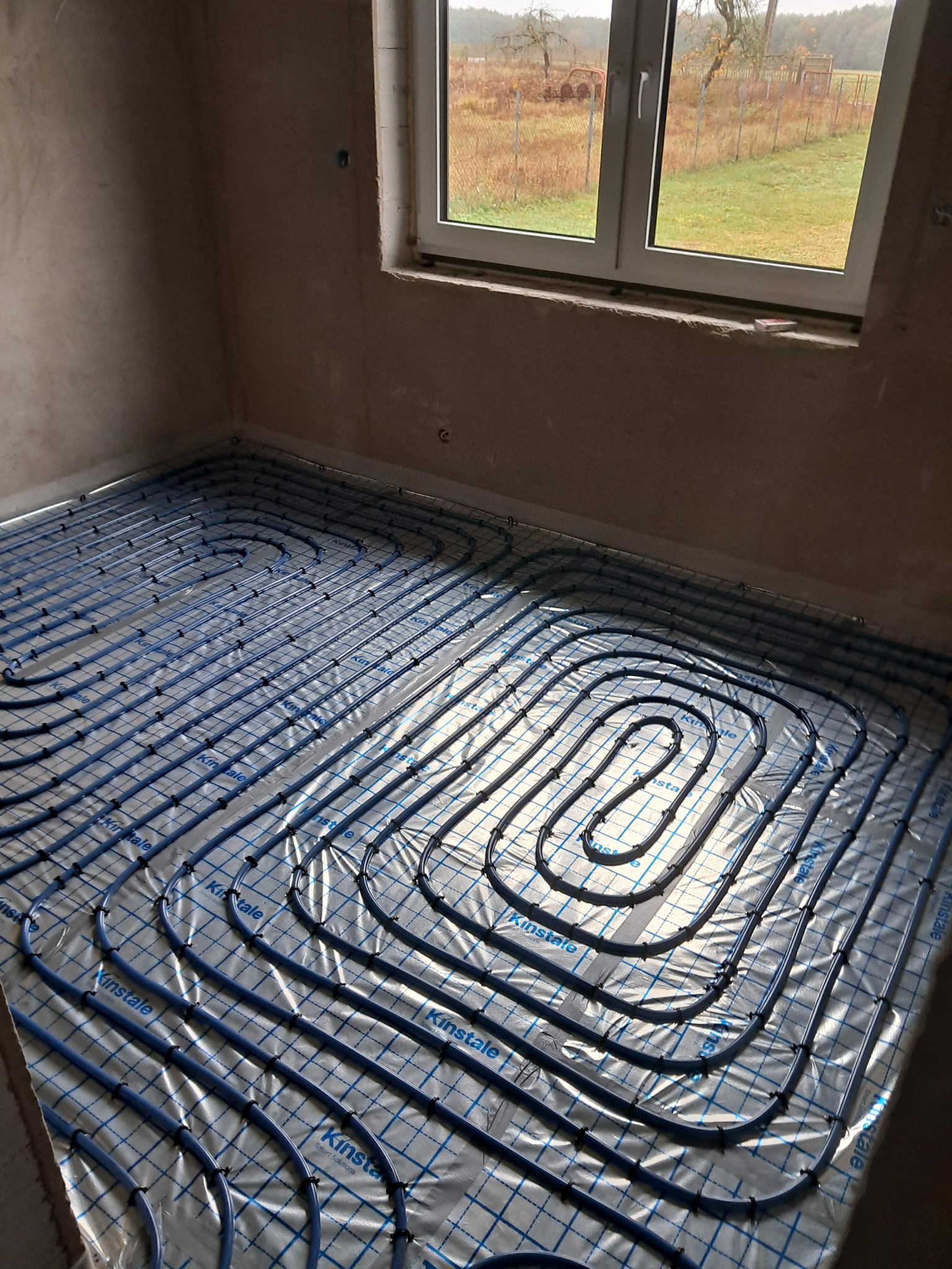 Niebieskie rury ogrzewania podłogowego ułożone na folii z nadrukiem 'Kinstale' w surowym wnętrzu z oknem, widok z góry.