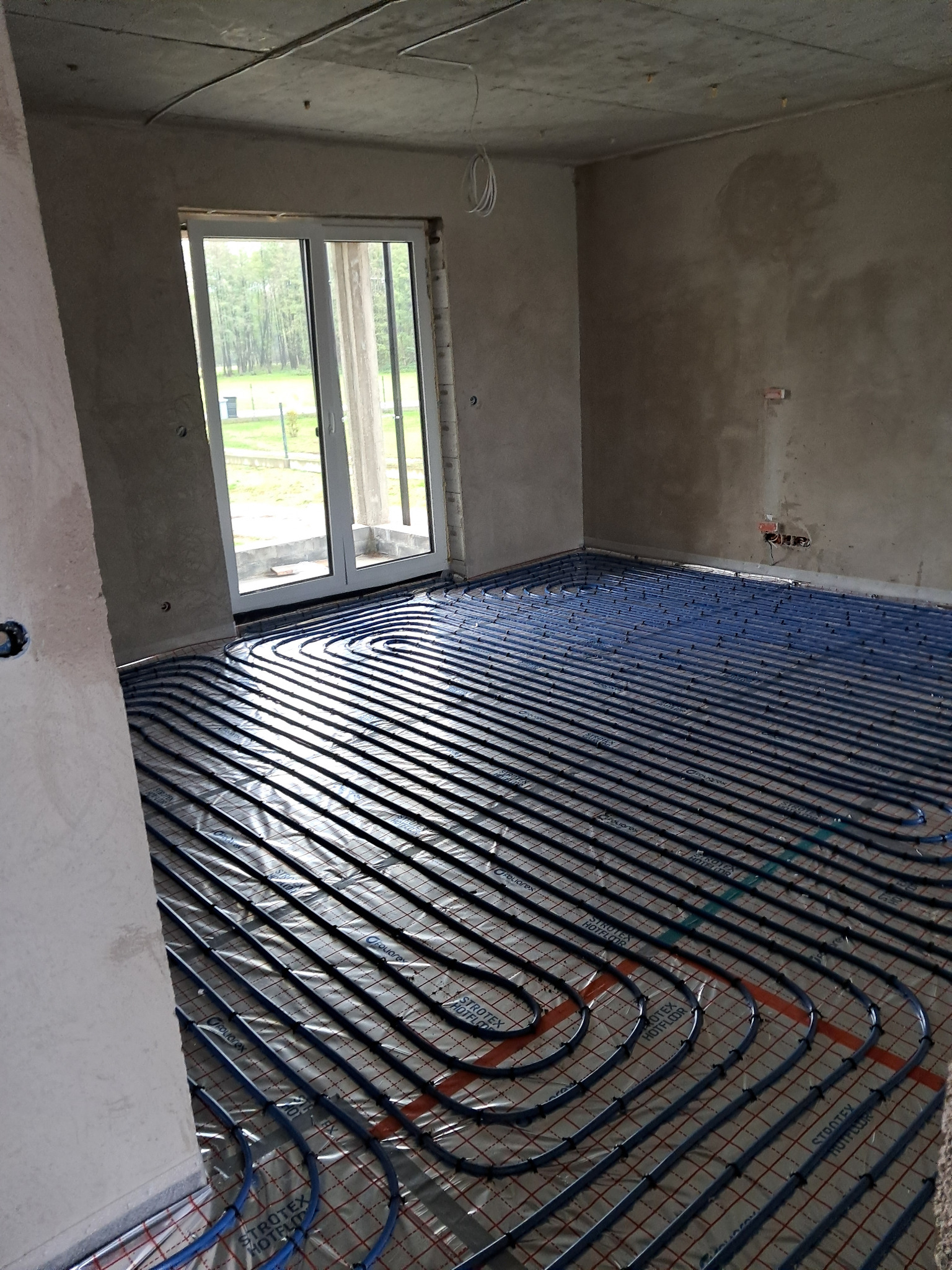 Niebieskie rury ogrzewania podłogowego ułożone na folii w surowym wnętrzu z betonowymi ścianami i wylewką, widoczne okno balkonowe.