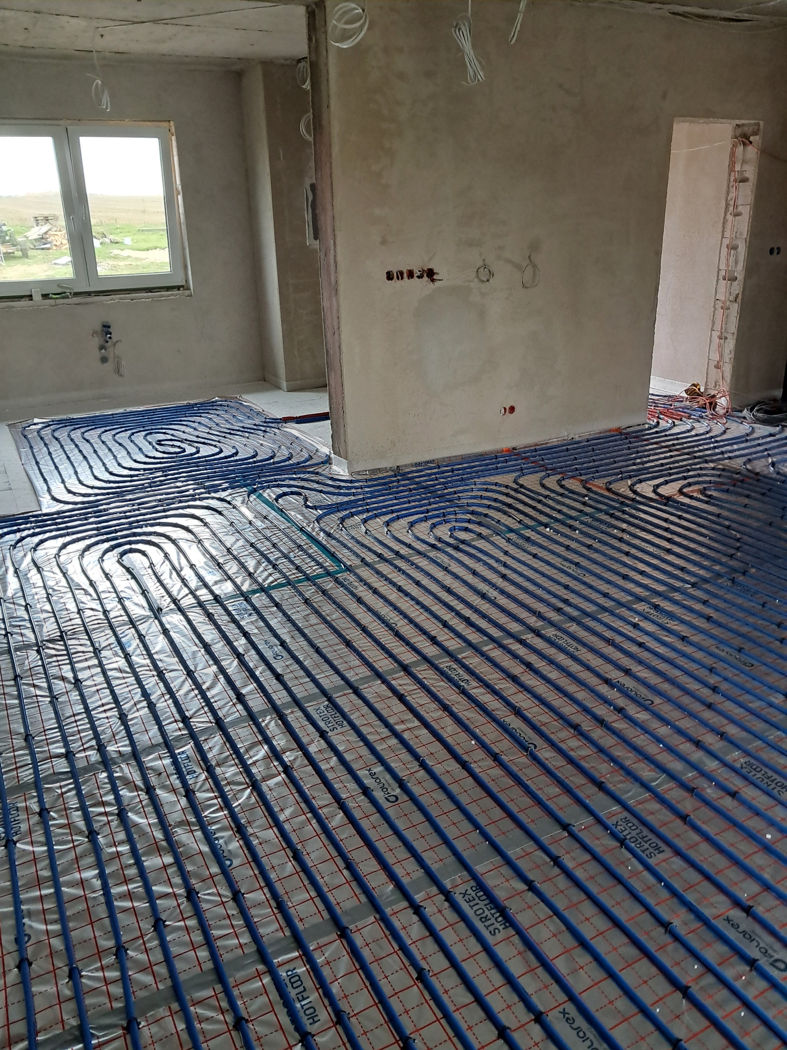 Niebieskie rury ogrzewania podłogowego ułożone na izolacji z siatką w surowym wnętrzu, widok z góry na dużą powierzchnię, okno w tle.