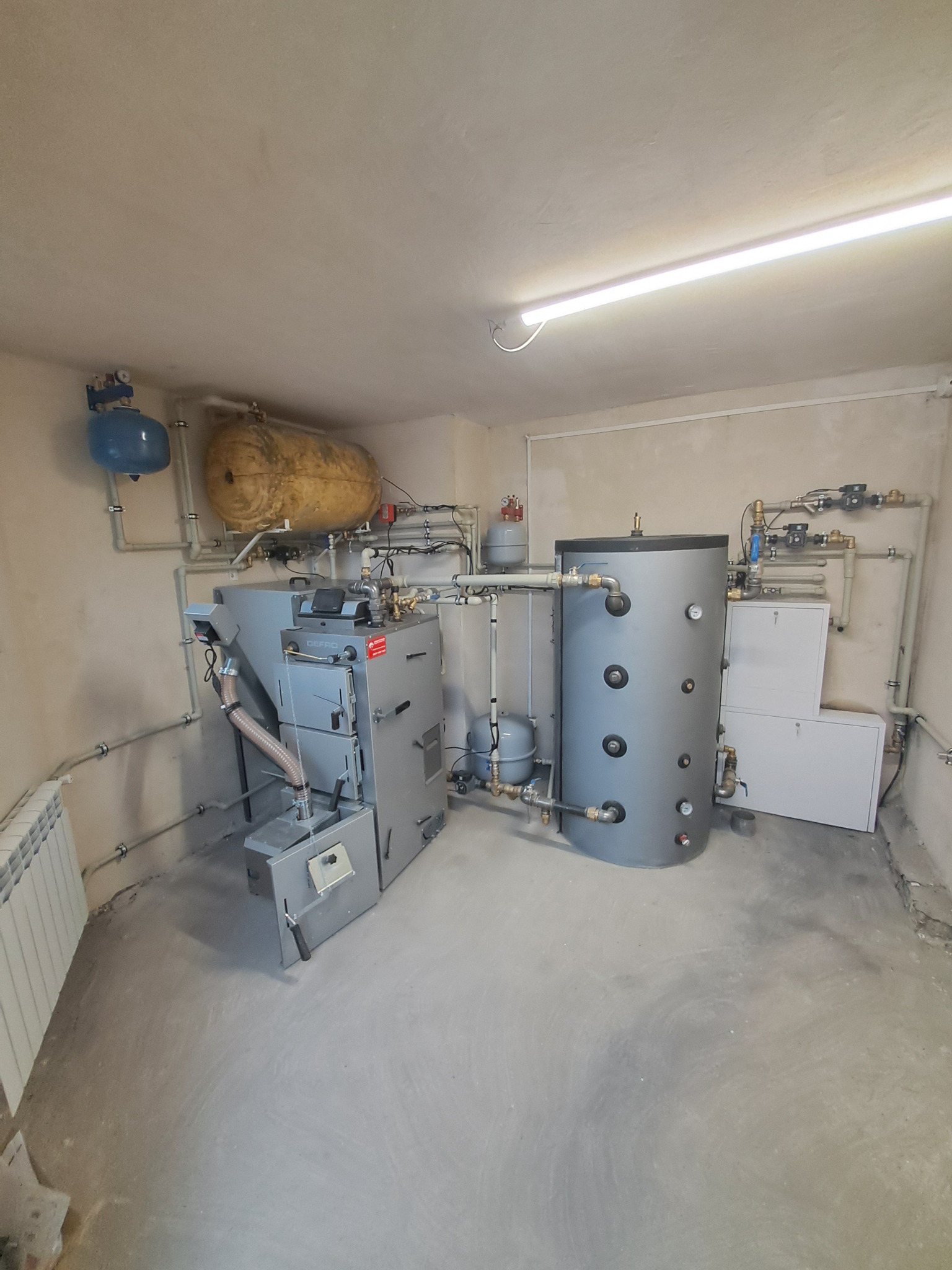 Kotłownia z piecem DEFRO na paliwo stałe, zasobnikiem ciepłej wody użytkowej, zbiornikiem wyrównawczym i orurowaniem w surowym pomieszczeniu.