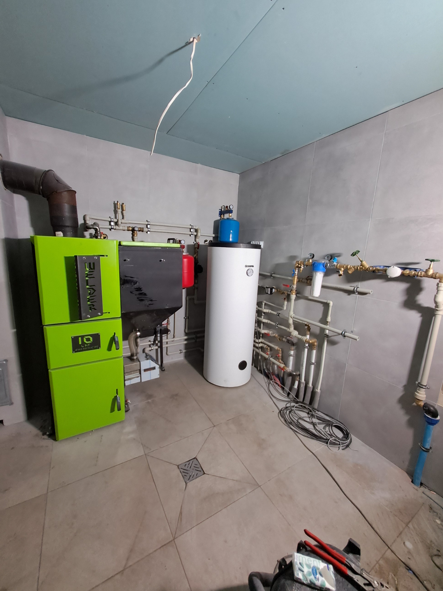 Kotłownia z nowoczesnym piecem na ekogroszek w odcieniach zieleni i czerni, białym zasobnikiem ciepłej wody, orurowaniem oraz narzędziami na podłodze. Widoczny fragment sufitu z wystającym kablem.