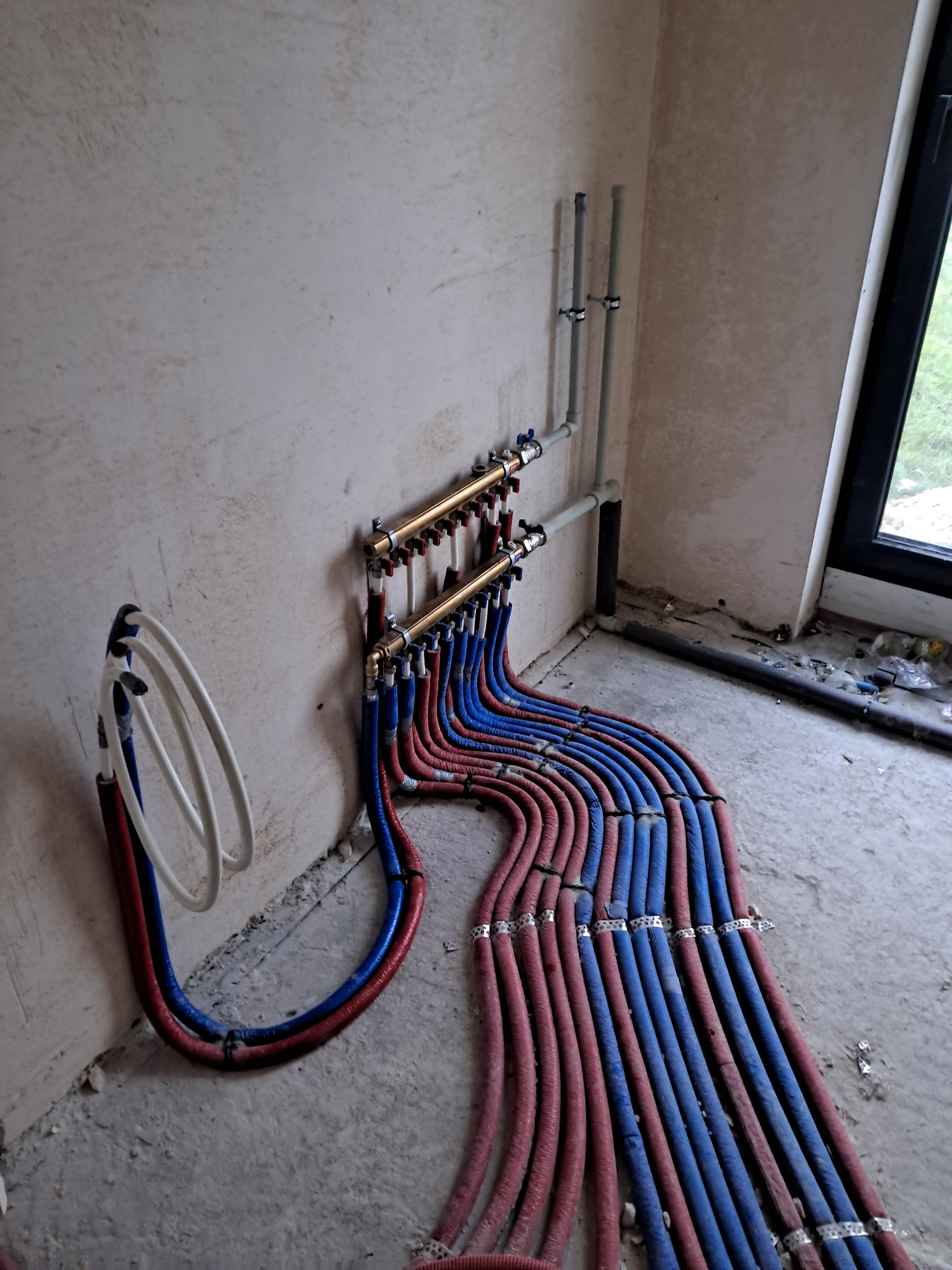 Rozdzielacz ogrzewania podłogowego z rurami PEX w kolorze niebieskim i czerwonym, zamocowany na ścianie w surowym wnętrzu, widoczne okno i elementy instalacji pionowej.