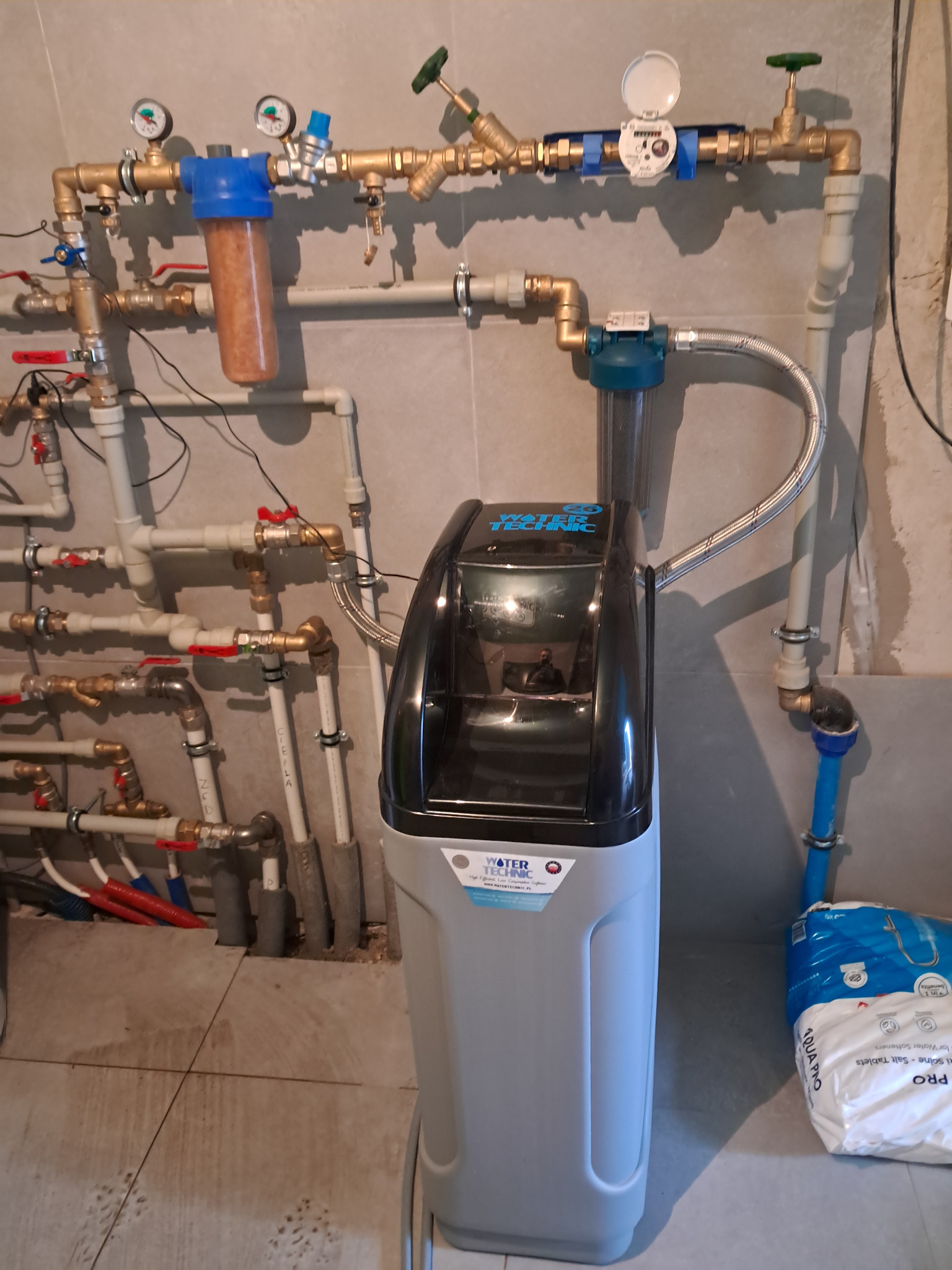 Zamontowany zmiękczacz wody Water Technic z licznymi rurami i filtrami, widoczny manometr i wodomierz, obok worek soli tabletkowanej Aqua Pro.